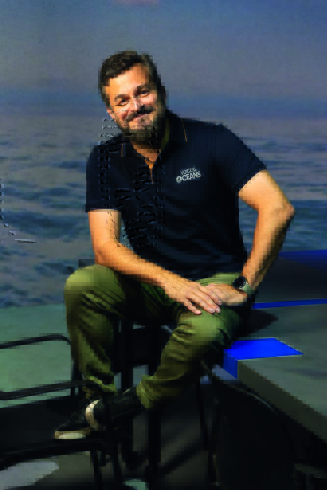 VOZESDOOCEANO13.jpg Homem grisalho posa sentado em mesa com polo azul e calça escura