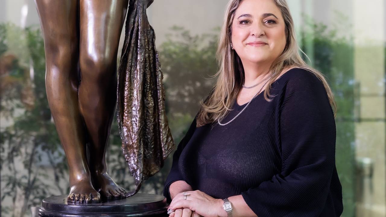 Moça loira posa com as mãos unidas ao lado de estátua. Veste peça preta.