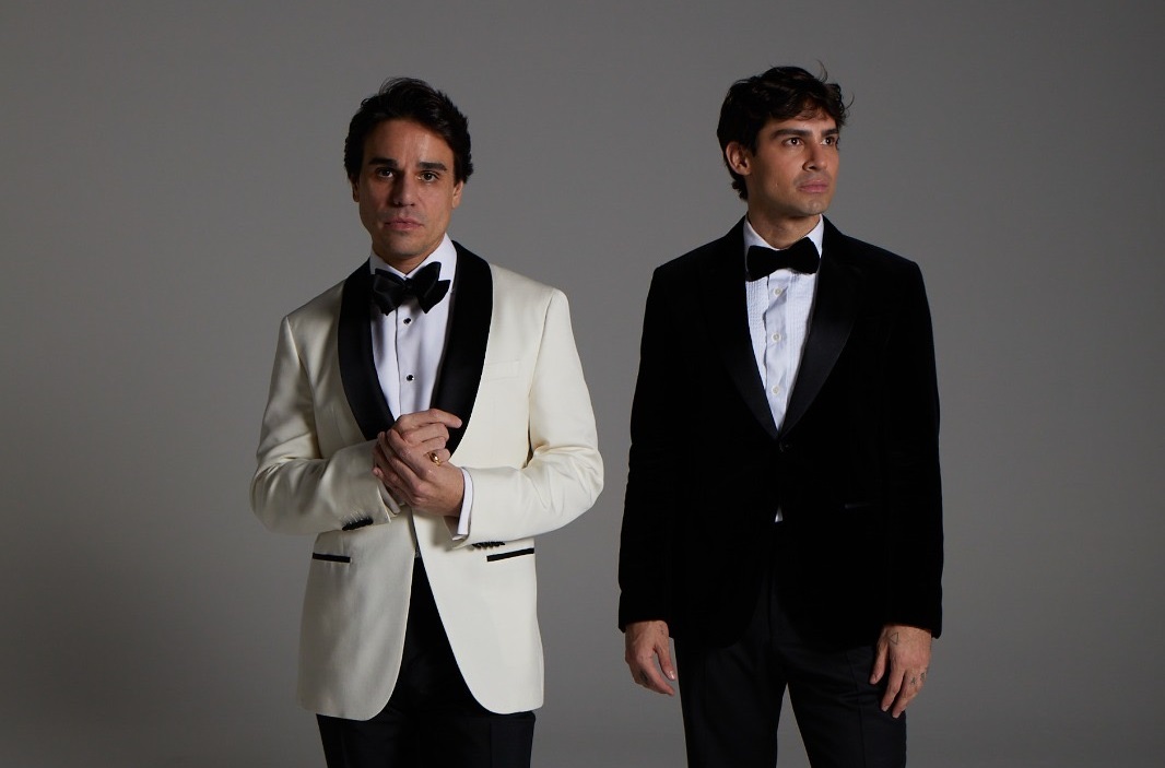Dois homens brancos posam de smoking, o da esquerda com paletó branco