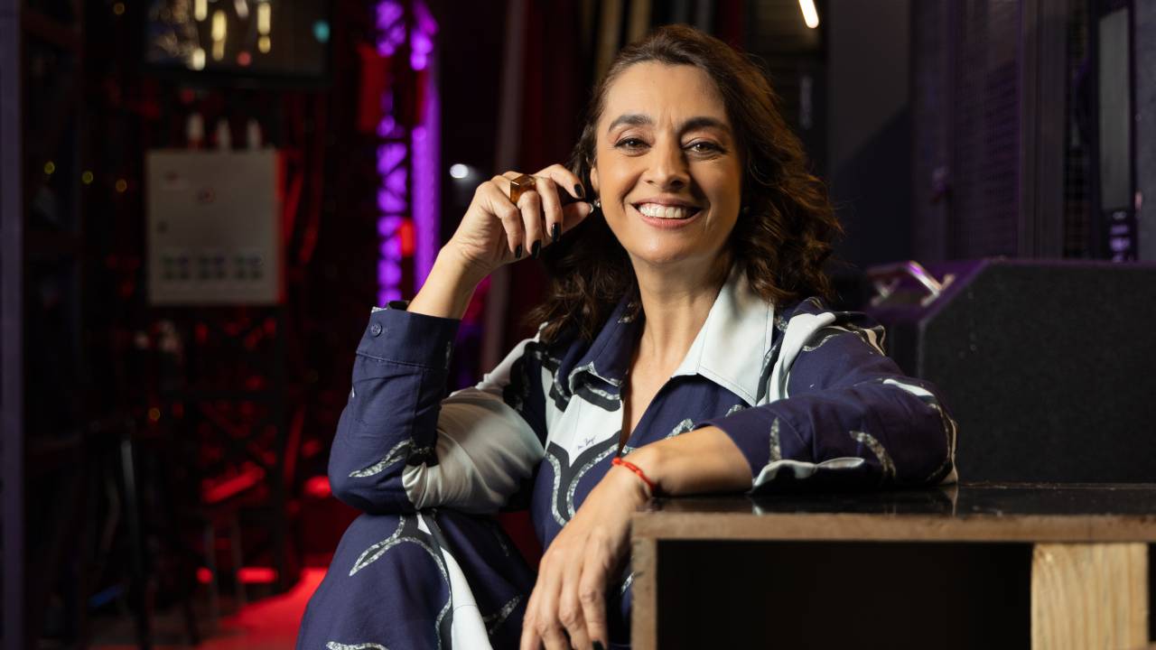 Mulher branca posa sentada com vestido azul e branco, sorri e coloca uma mão no rosto