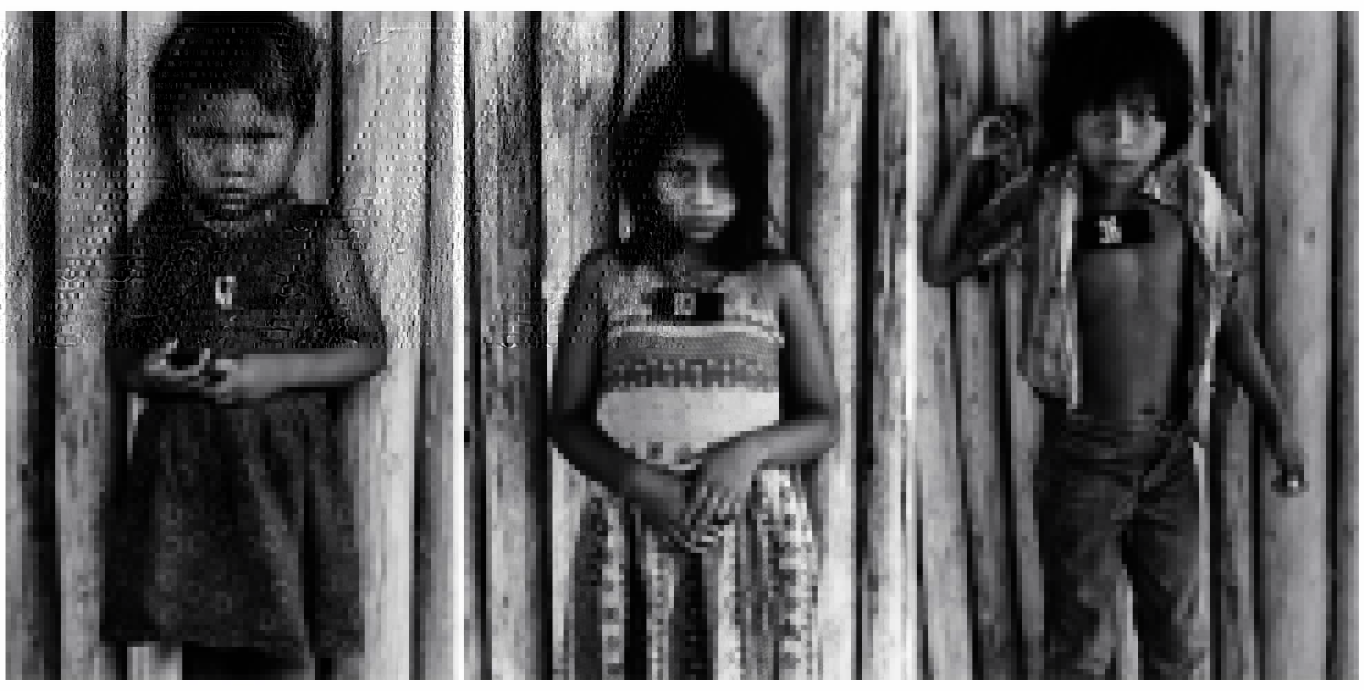 Fotografia de três crianças indígenas encostadas em área de madeira