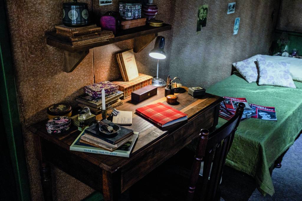 Foto de mesa com diário, luminária e cama ao fundo