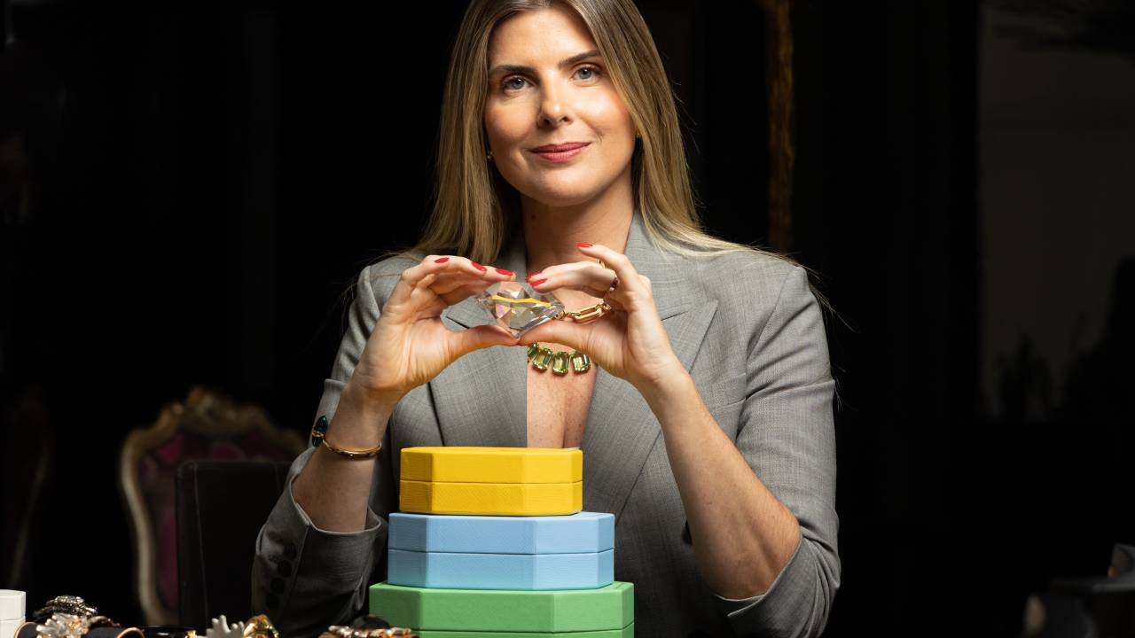 Mulher loira de blazer cinza posa segurando grande cristal apoiada em mesa preta com caixas verde, azul e amarelo e lotada de joias