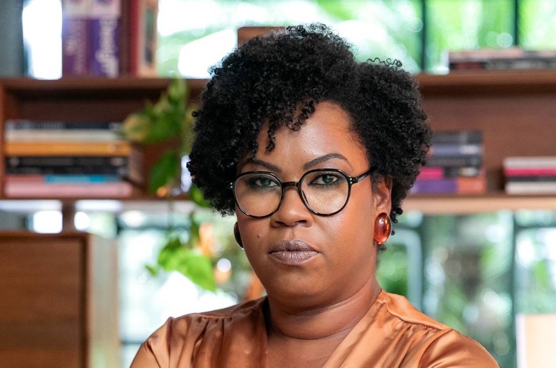 Mulher negra de óculos redondos posa sentada vestindo peça de seda em tom marrom e calça branca