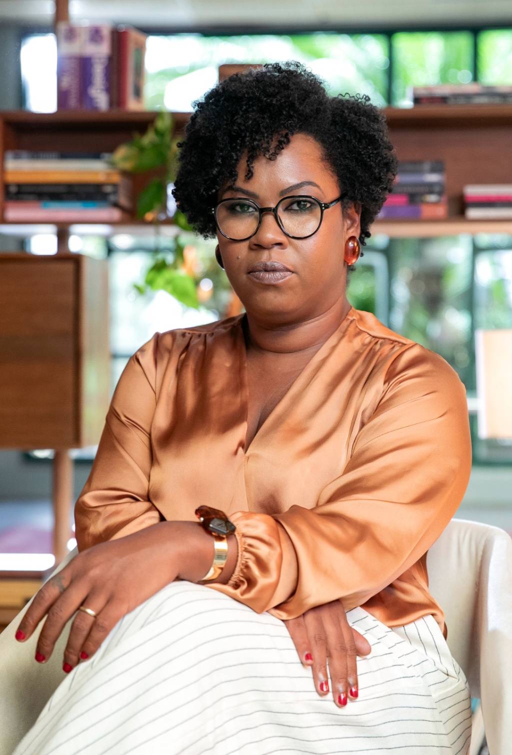 Fayda Belo Crédito Manoella Mello-Divulgação Mulher negra de óculos redondos posa sentada vestindo peça de seda em tom marrom e calça branca