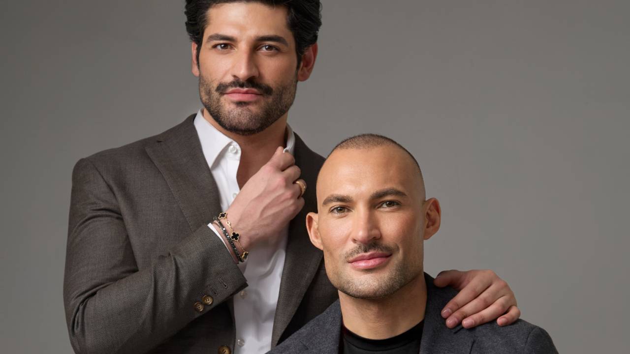 Homem de barba e cabelos negros posa de pé ao lado de homem branco e careca, ambos sorrindo e vestindo ternos escuros.
