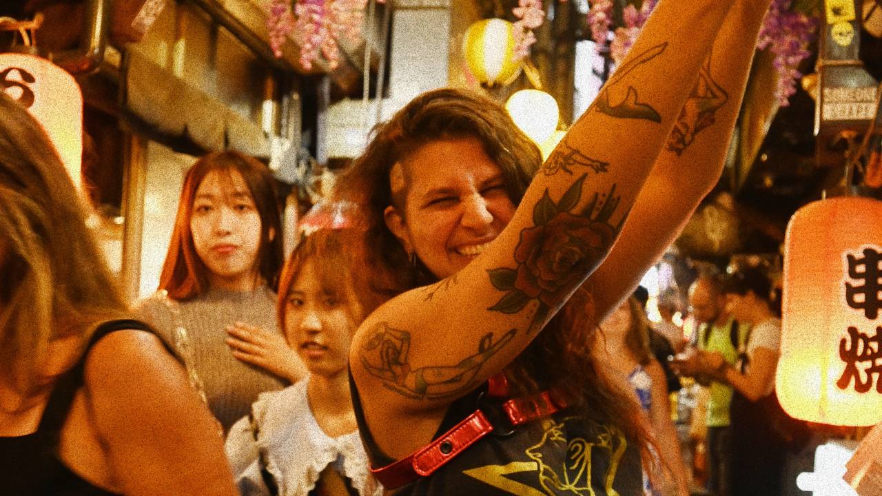 Mulher branca de cabelos encaracolados longos posa rindo e segurando lâmpada japonesa em corredor estreito de turistas