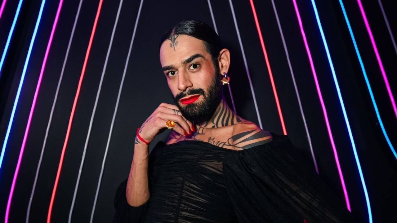 Travesti de barba e batom vermelho posa em frente a neon com mão no queixo e roupa preta.