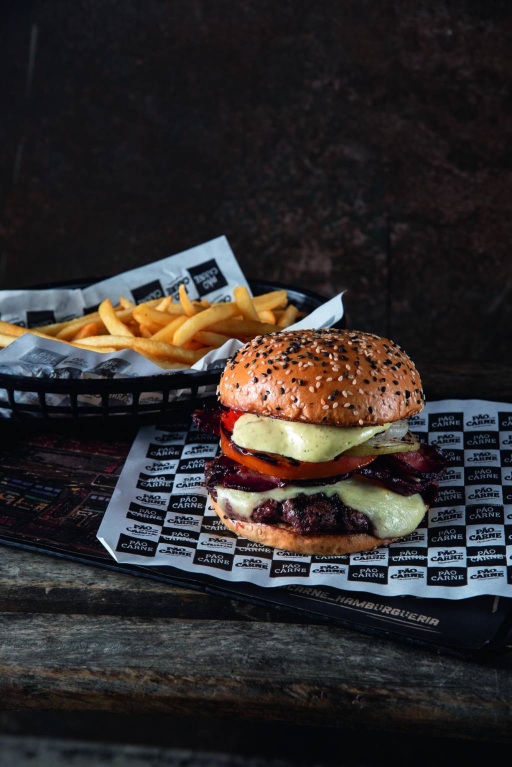Comer & Beber 2024 – Pão com Carne Hambúrguer em cima de papel com logo da lanchonete e porção de batata frita ao fundo