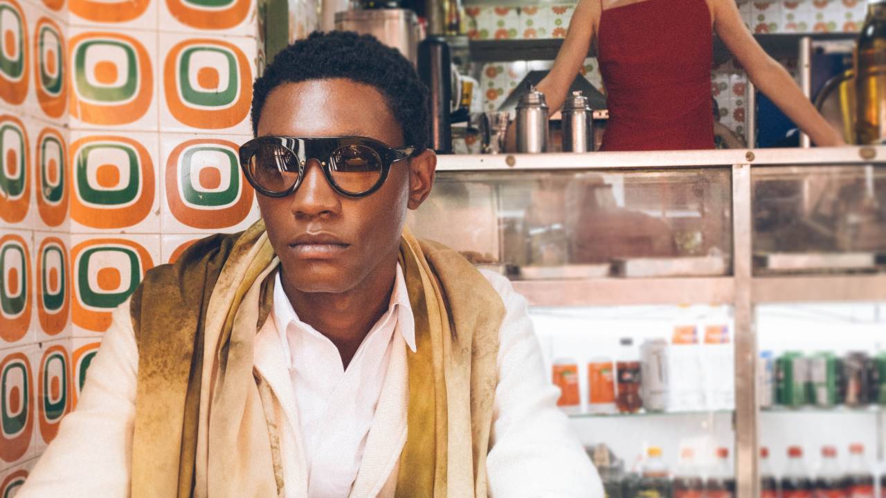 Moço negro de óculos e peça branca sentado em primeiro plano em mesa de bar. No balcão, moça branca de vestido vermelho aparece de pé