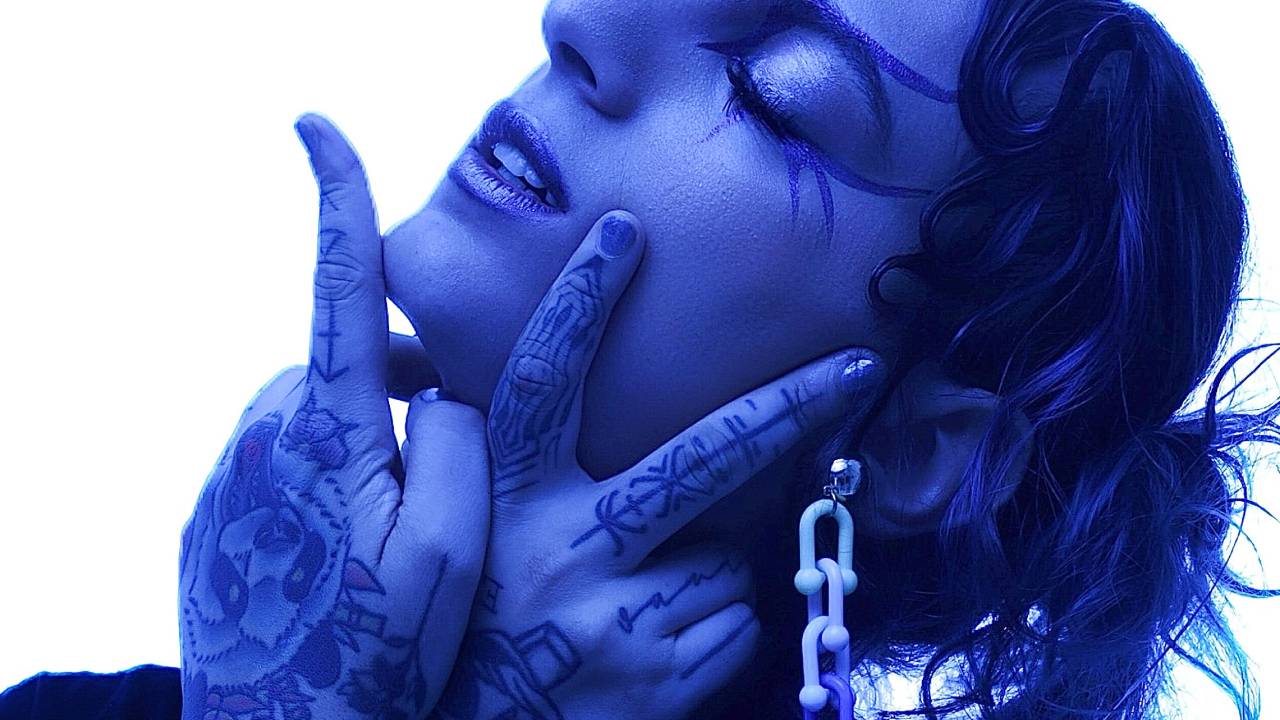 Moça iluminada em tom azul posa com o rosto voltado pra cima, olhos fechados, as mãos com os dedos exibindo "dois" e tatuagens