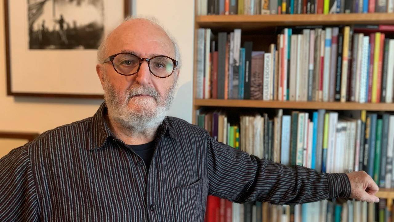 Senhor de barba grisalha e óculos posa apoiado ao lado de estante de livro, vestindo camisa preta listrada fechada