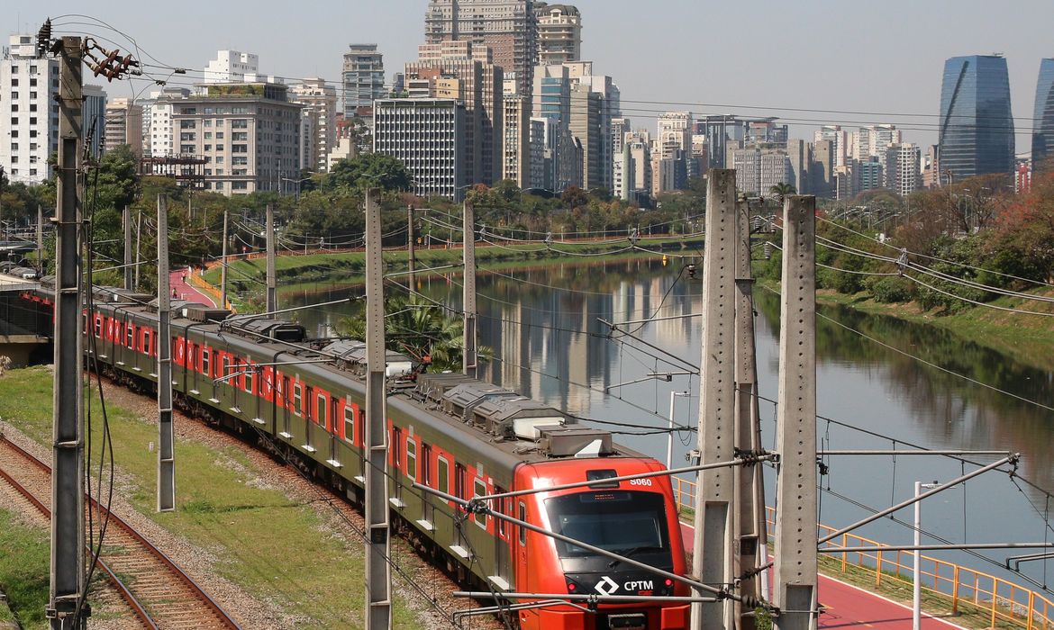Trem passando nos trilhos com rio e prédios ao fundo