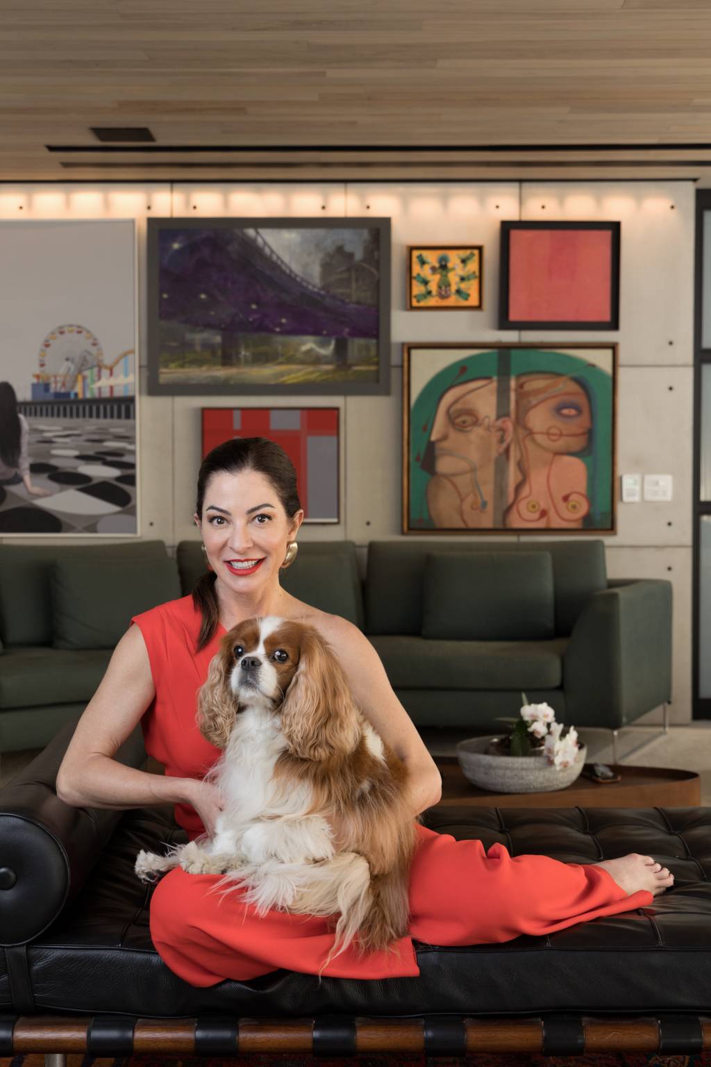 Ana Paula Padrão Crédito Wanezza Soares-VejaSP (02) Mulher branca de peça alaranjada posa com cachorro branco e marrom no colo em divã de couro preto e galeria de quadros e sofá verde ao fundo