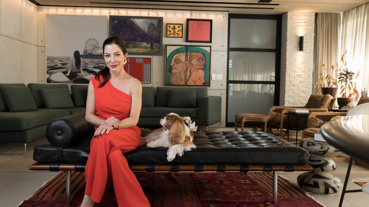 Mulher branca de peça alaranjada posa com cachorro branco e marrom no colo em divã de couro preto e galeria de quadros e sofá verde ao fundo