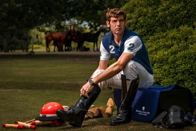 Homem com roupa azul e branca do esporte polo, sentado na grama com capacete laranja aos pés
