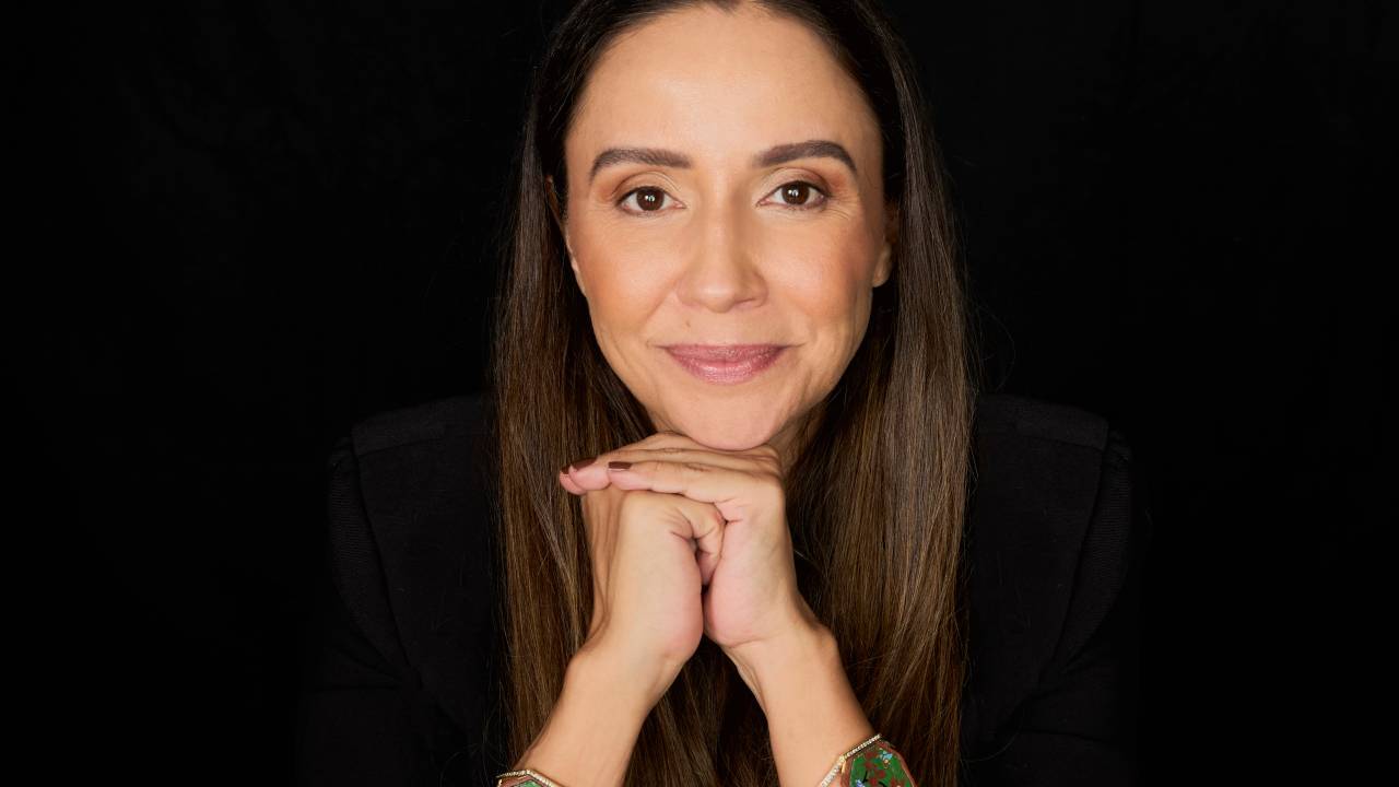 Mulher branca de cabelos longos com mãos no queixo sentada em mesa com pulseiras à mostra