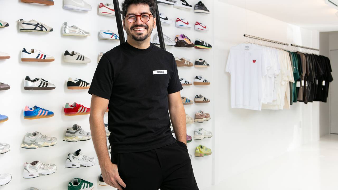 Lucas Cimino, da Zipper Galeria, em nova loja dedicada a peças exclusivas de steetwear. Ele veste peças pretas e óculos de grau em frente a parede com vários tênis alinhados.