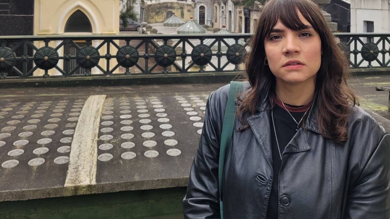 Mulher de franja e cabelo longo com jaqueta em local que aparenta ser um cemitério antigo