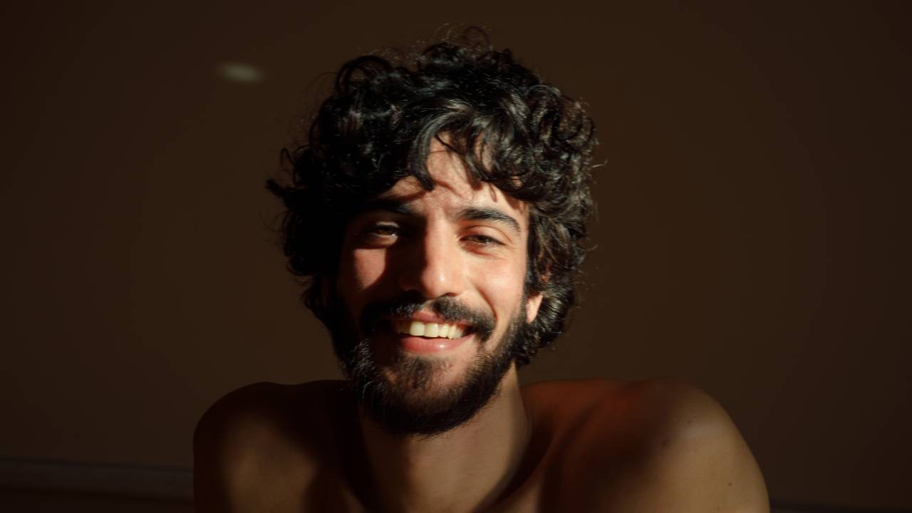 Moço de cabelo preto encaracolado posa sorrindo sem camisa, com a luz batendo no rosto
