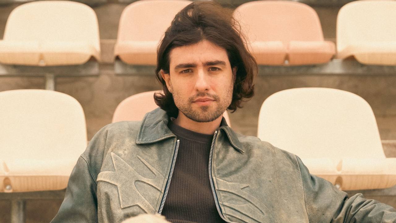 Homem de cabelos ligeiramente longos posa sentado em cadeira de estádio