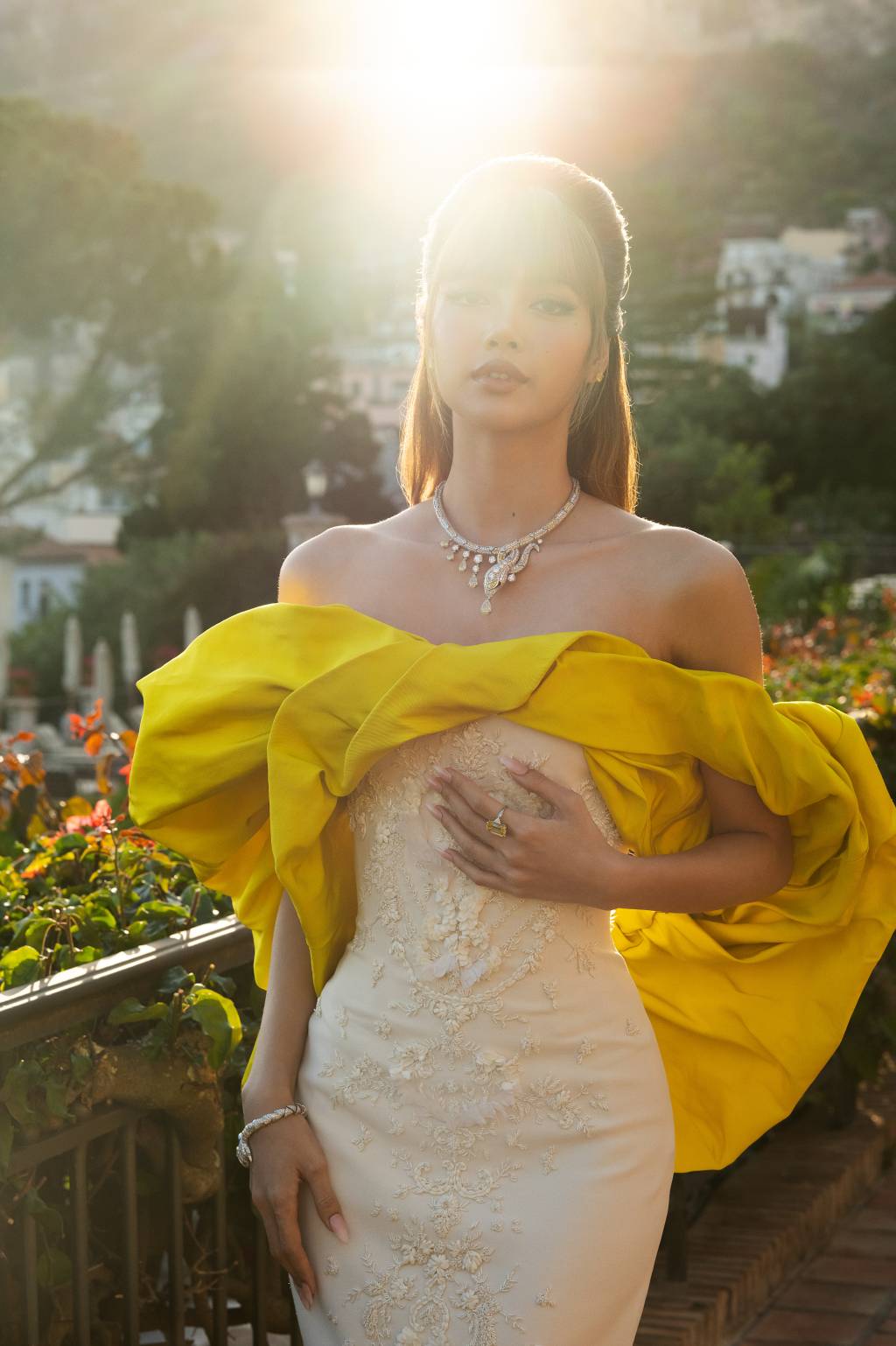 2025.05.19 Cannes – Bvlgari Lisa L1011944 RT2 A atriz e cantora sul-coreana Lisa Manobal em vestido branco com peça amarela ao redor do colo vestindo um colar dourado em jardim com sol batendo ao fundo