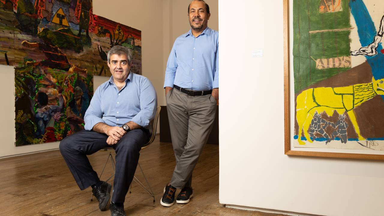 Os galeristas Carlos e Antônio, ambos de camisa azul claro, Carlos sentado e Antônio encostado na quina da parede com quadro colorido à direita