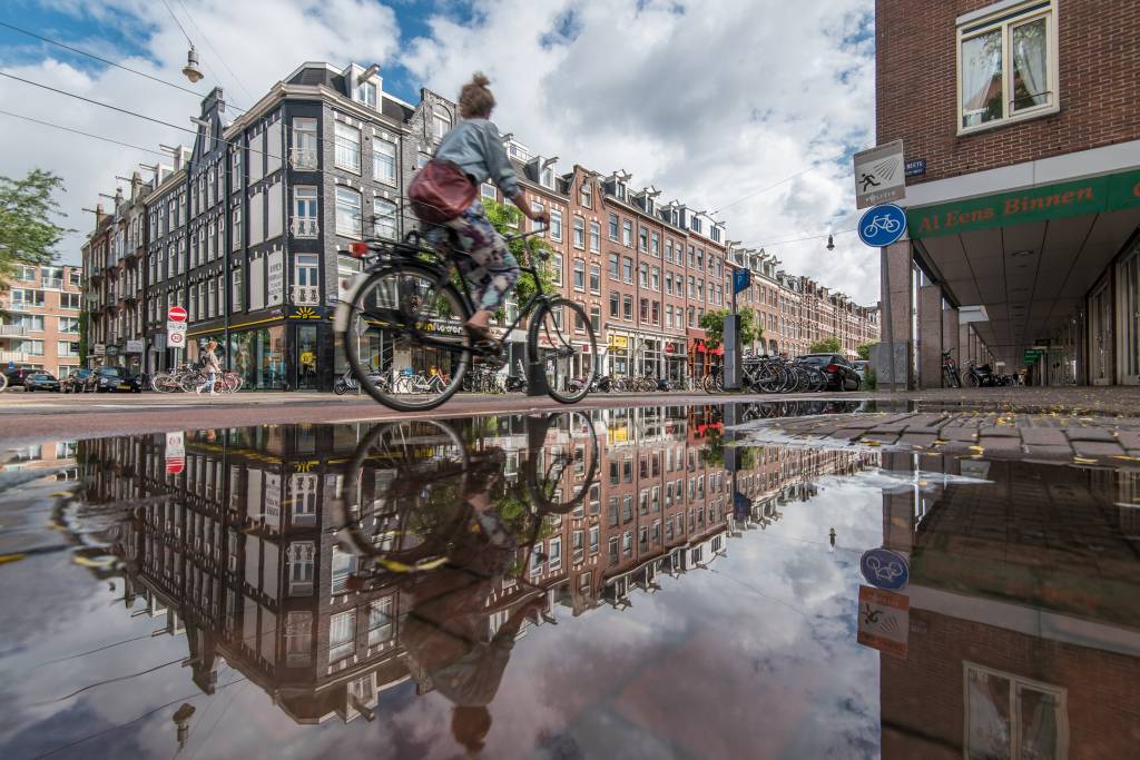 Cycling Kinkerstraat -Koen Smilde Photography-Rechtenvrij Crédito Koen Smilde Photography Foto do reflexo de uma poça mostrando moça de bike passando por Amsterdã