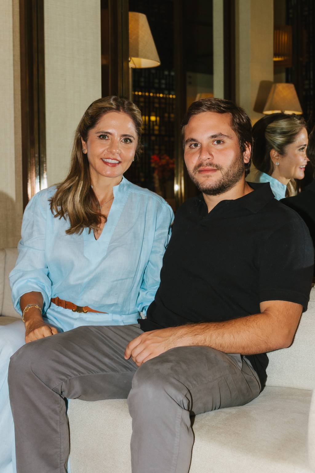 Flavia e João Paulo Nasser Crédito Daniel Araújo-Divulgação Mãe e filho sentados em sofá, ela com camisa de seda azul claro, ele de polo escura