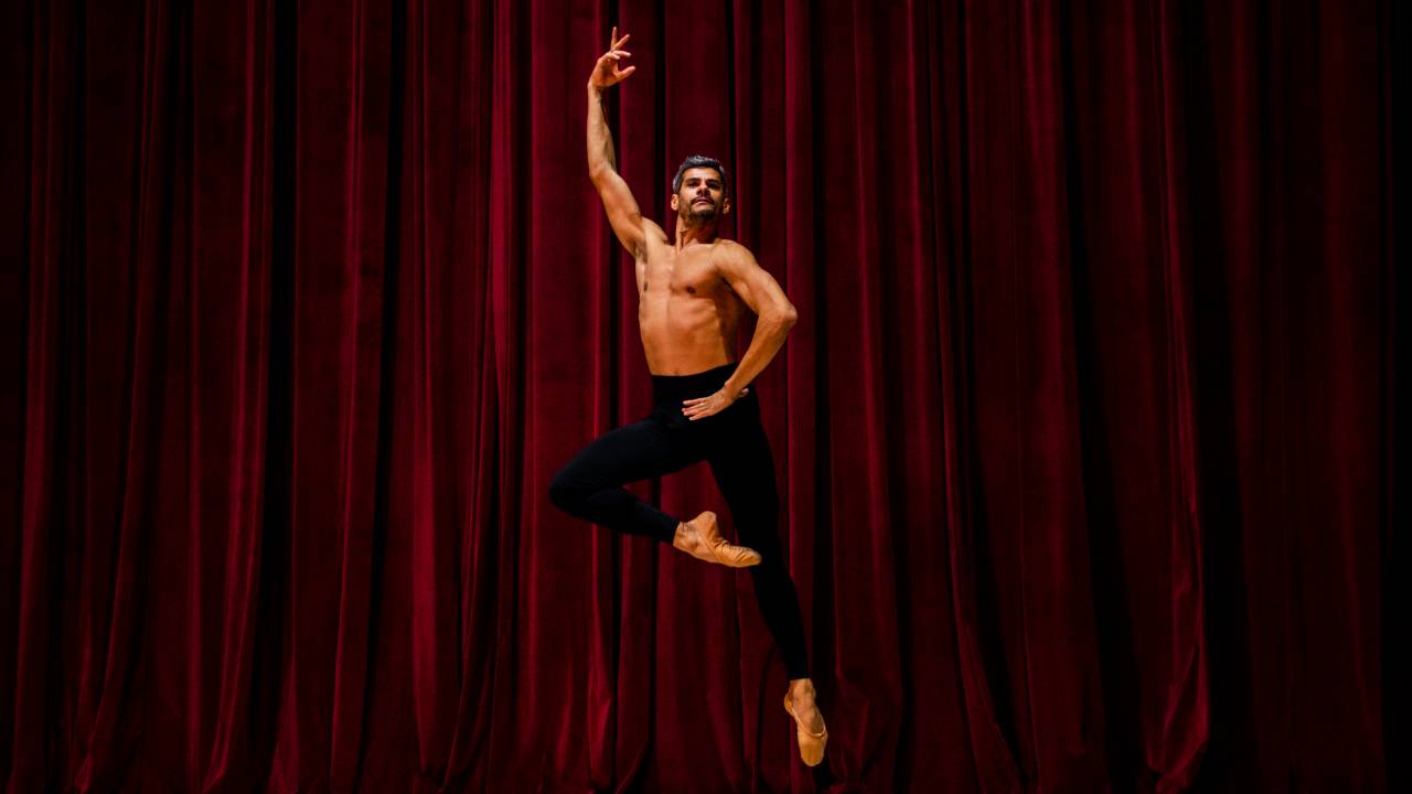 Thiago Soares pulando em pose de dança em frente a cortina vermelha