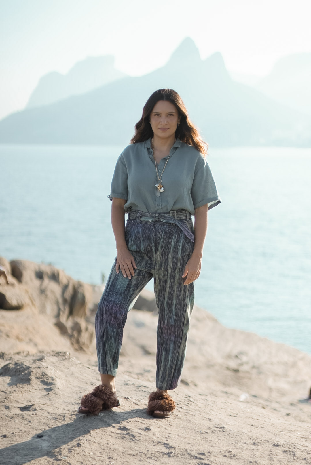 Mulher em penhasco com mar ao fundo, vestindo pe&ccedil;as azuis e cinza