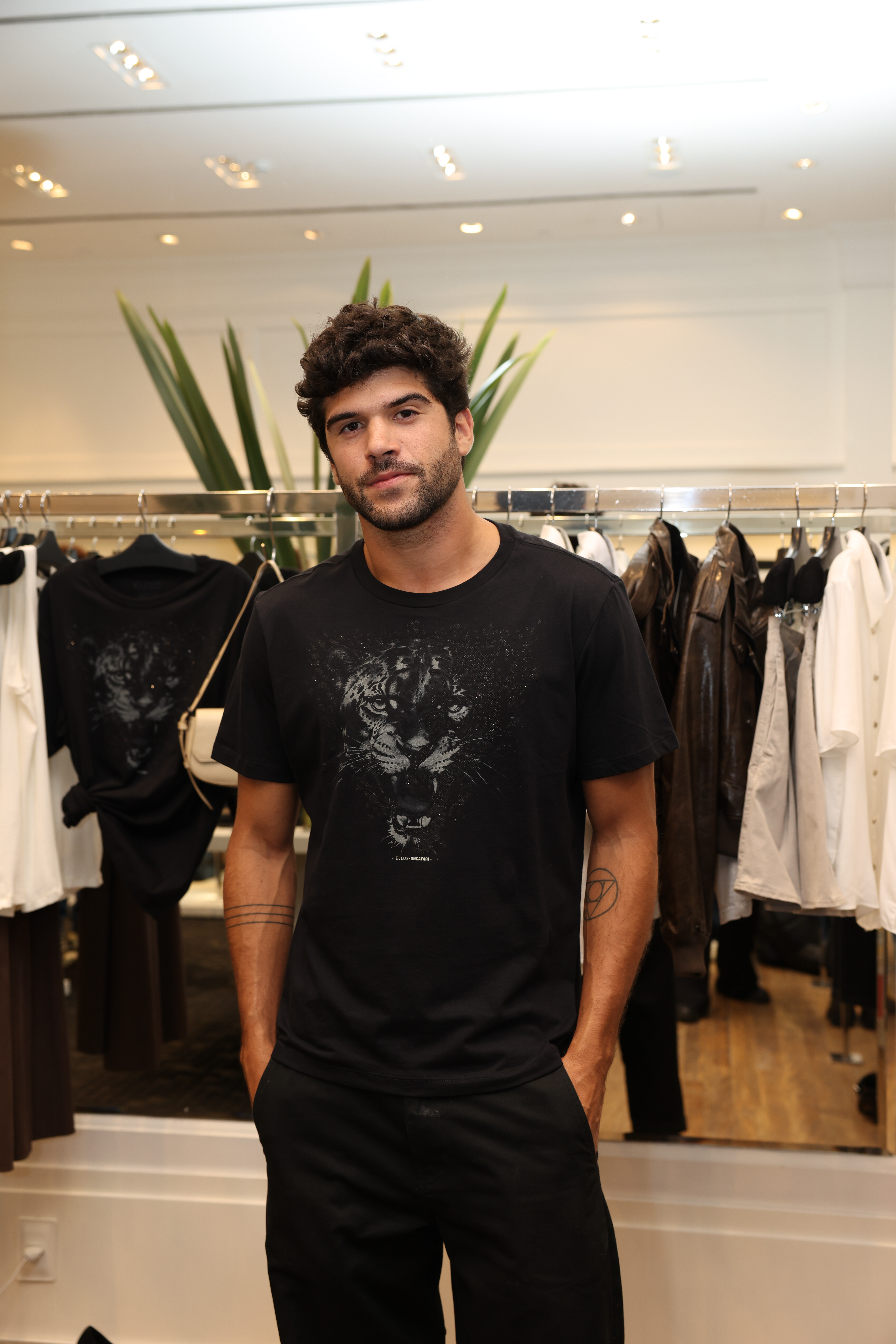 Homem branco de barba posa com roupas pretas em loja de roupas