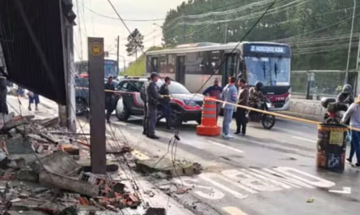 Cena de acidente com ônibus e carro de polícia em rua com escombros, cones e fita de isolamento. Pessoas e motocicletas observam a situação.