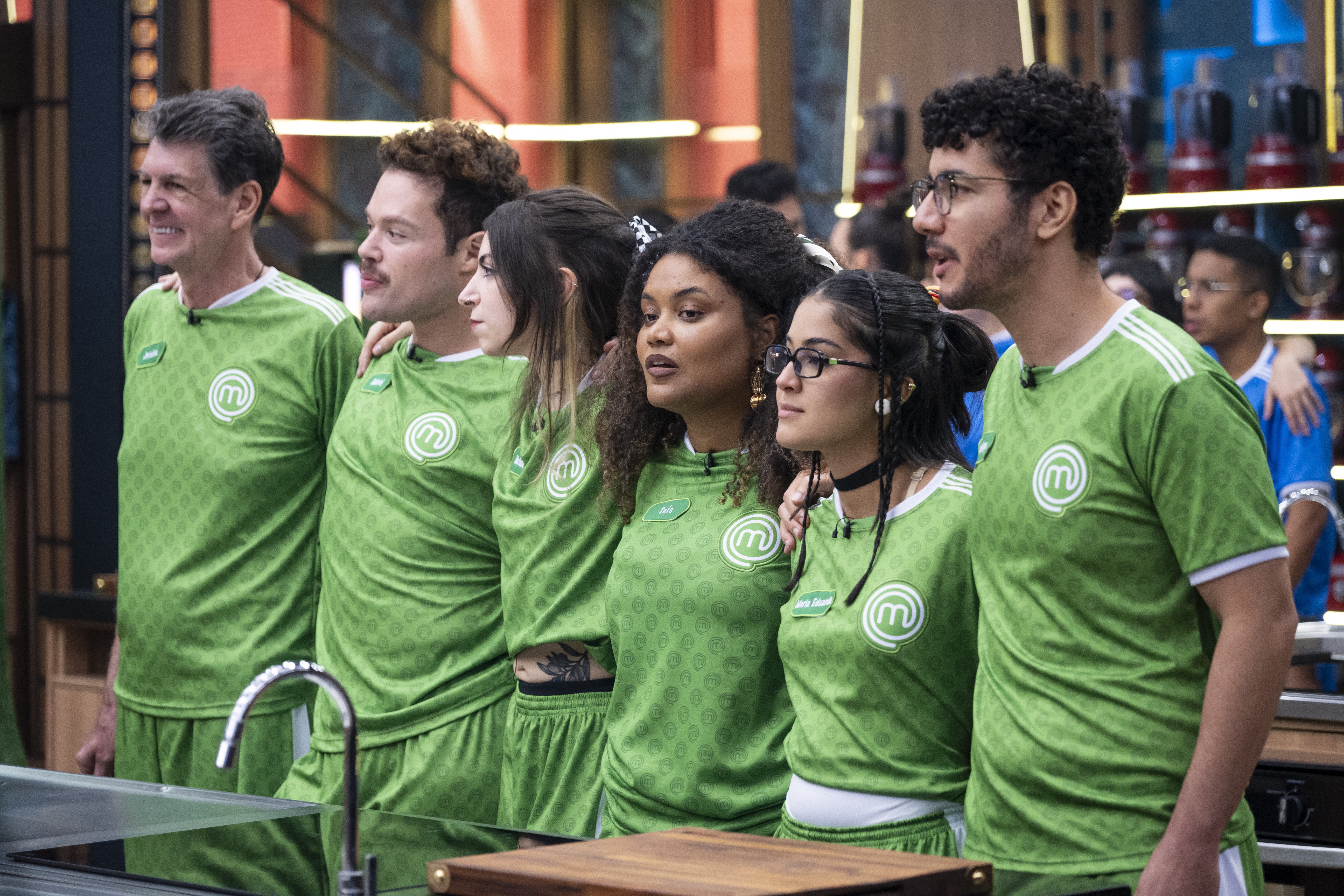 Seis participantes do MasterChef Brasil, três homens e três mulheres, em pé, lado a lado, vestindo uniformes verdes com o logo do programa. Eles olham para a direita, com expressões atentas, em uma cozinha profissional