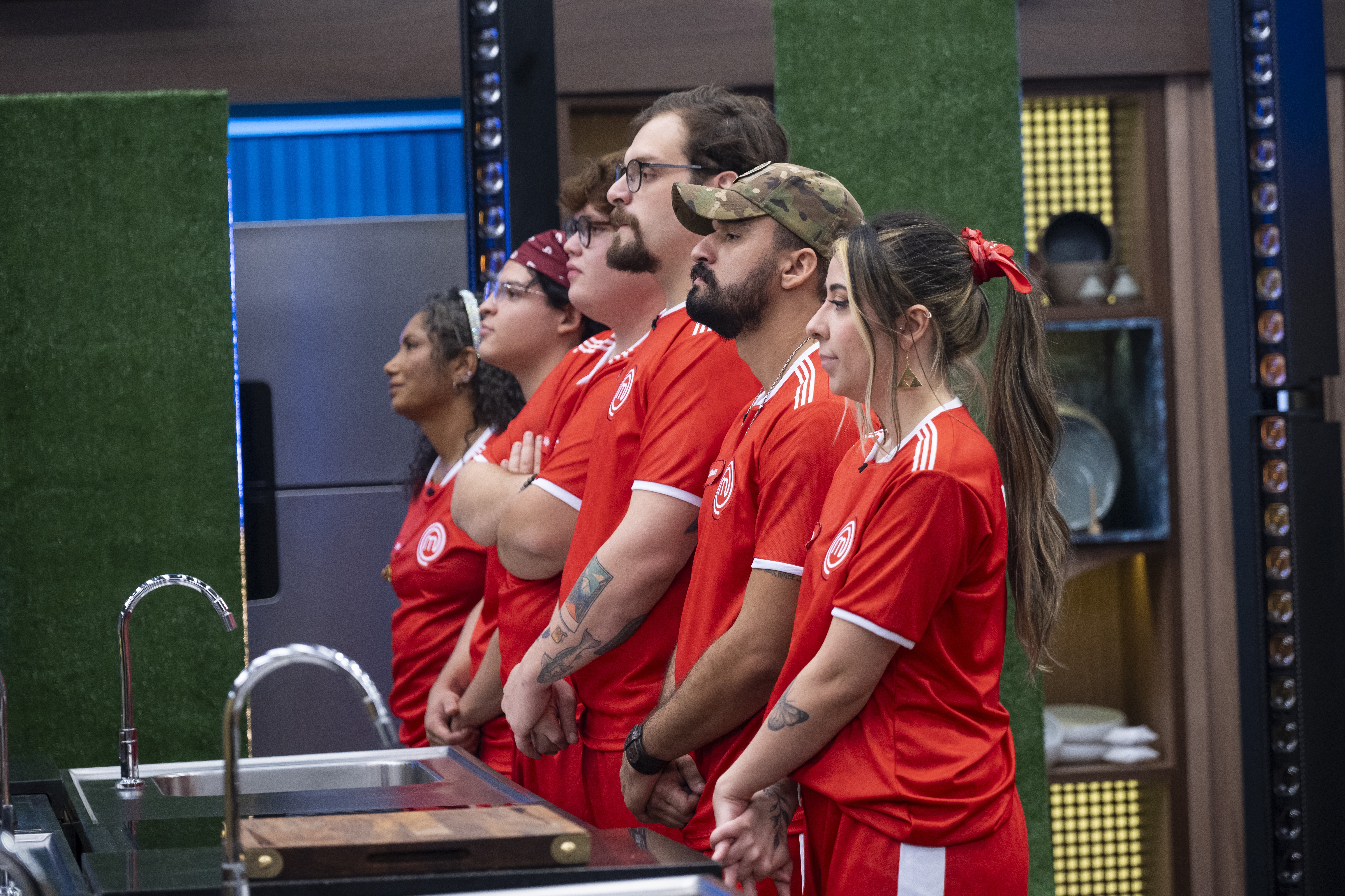 Cinco pessoas, três homens e duas mulheres, em fila, vestindo uniformes de futebol vermelhos com listras brancas, observam algo à esquerda. Eles estão em uma cozinha com pias de inox e uma tábua de corte de madeira em primeiro plano