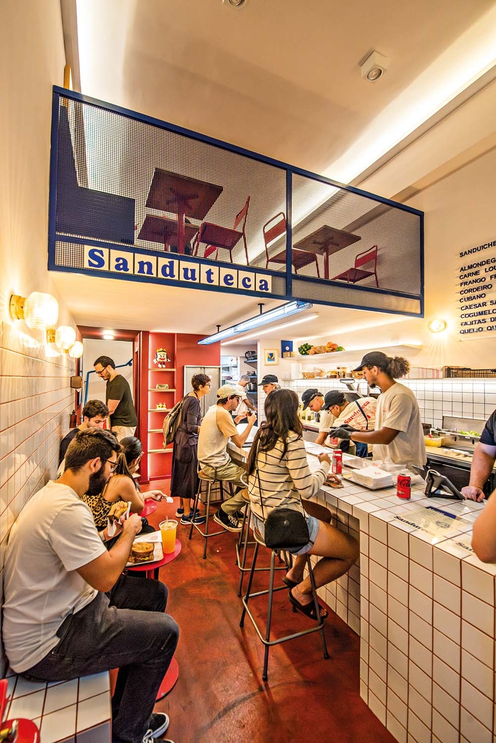 Interior de uma lanchonete moderna com piso vermelho e paredes de azulejos brancos. Clientes comem em mesas e balcões, enquanto funcionários preparam lanches. Um letreiro azul com a palavra 