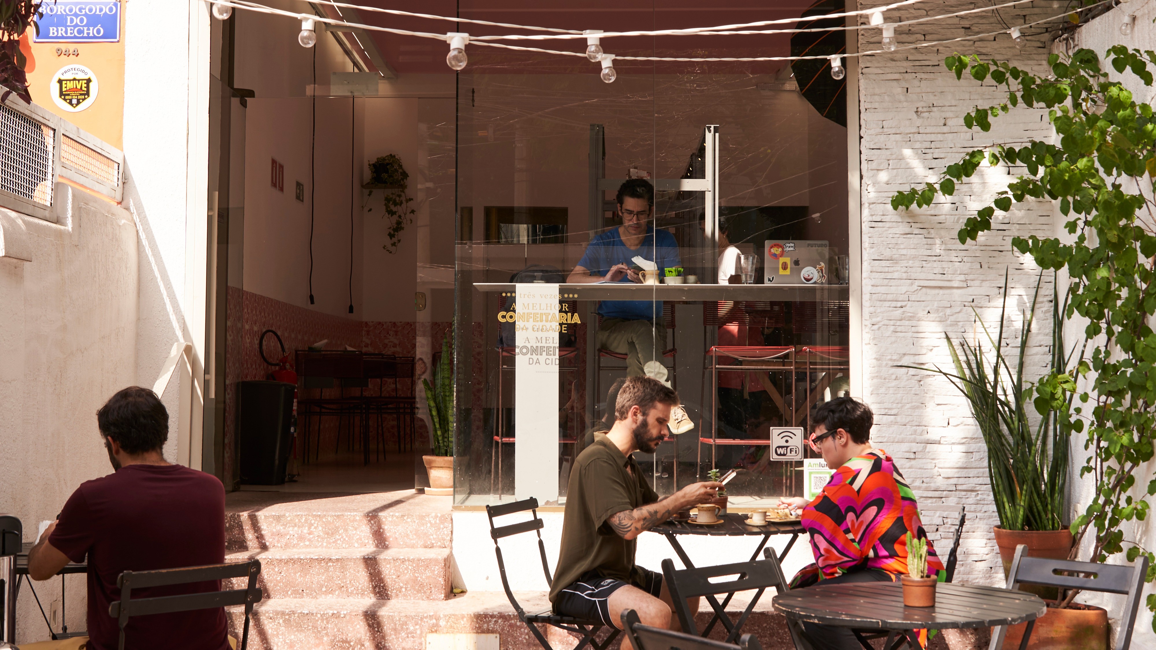 Três pessoas em um café: um homem de barba e camiseta verde-oliva usa o celular em mesa externa, uma mulher de óculos e blusa colorida senta à frente, e outro homem de camiseta azul trabalha no balcão interno. Plantas decoram o ambiente externo com luz solar
