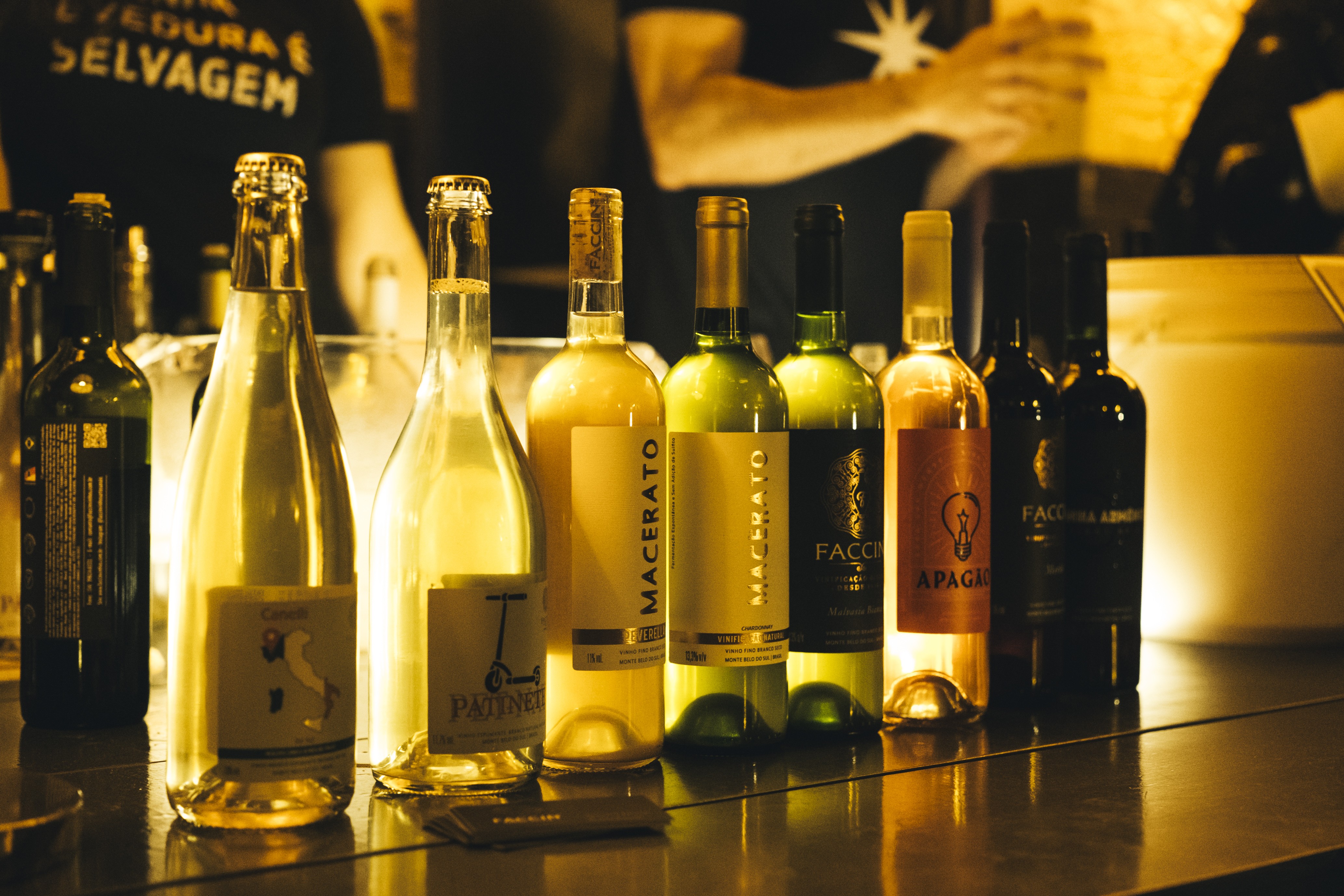 Garrafas de vinho e espumante em tons de verde, âmbar e transparente, alinhadas sobre um balcão escuro e brilhante, com iluminação quente ao fundo, onde uma pessoa de camiseta preta com a frase SELVA E SELVAGEM é visível