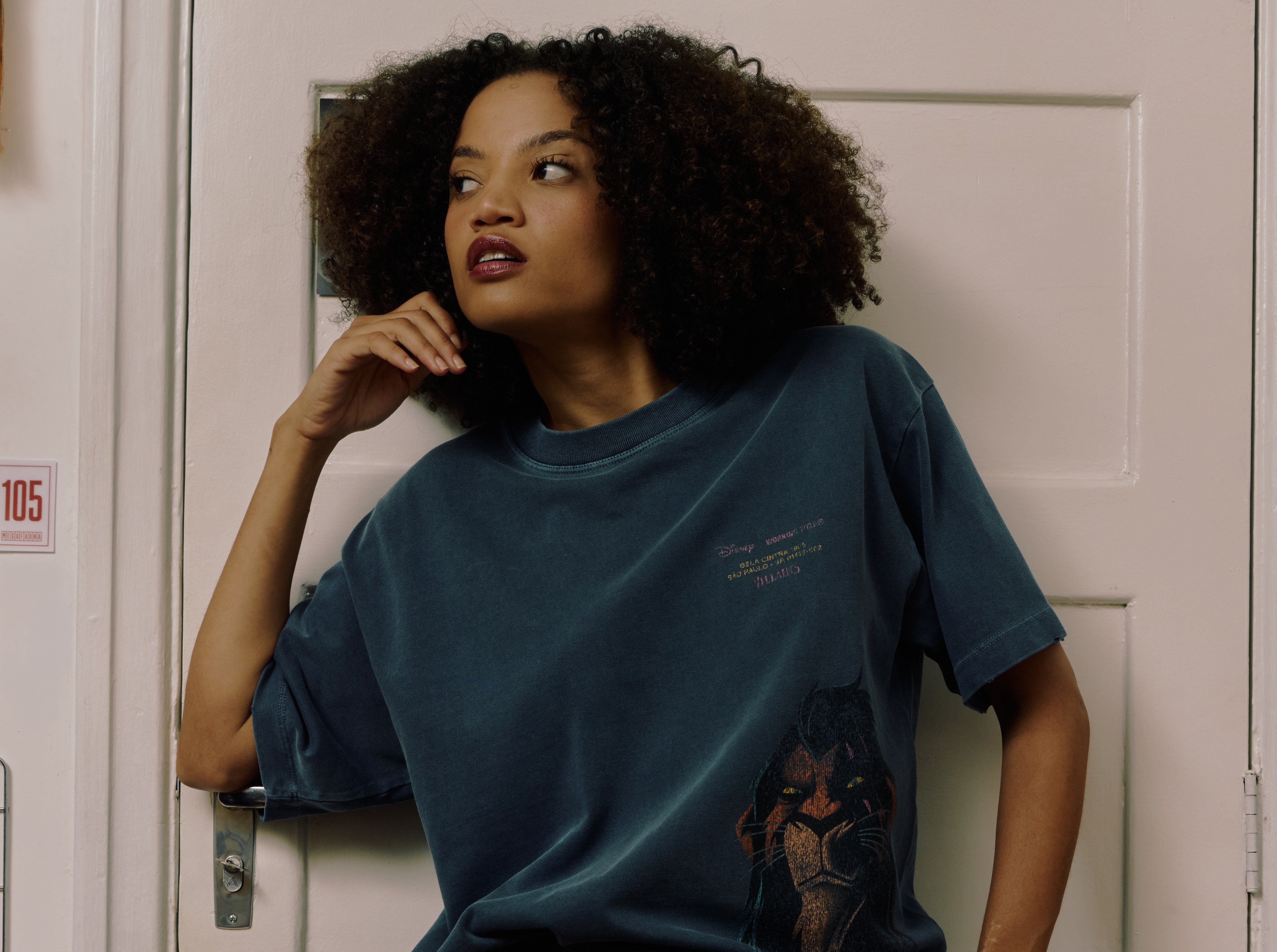 Mulher jovem de pele morena e cabelo cacheado escuro, vestindo uma camiseta azul-petróleo com estampa do Scar, personagem de O Rei Leão, e um pequeno texto. Ela está com a mão direita no queixo, olhando para a direita, em frente a uma porta branca com o número 105