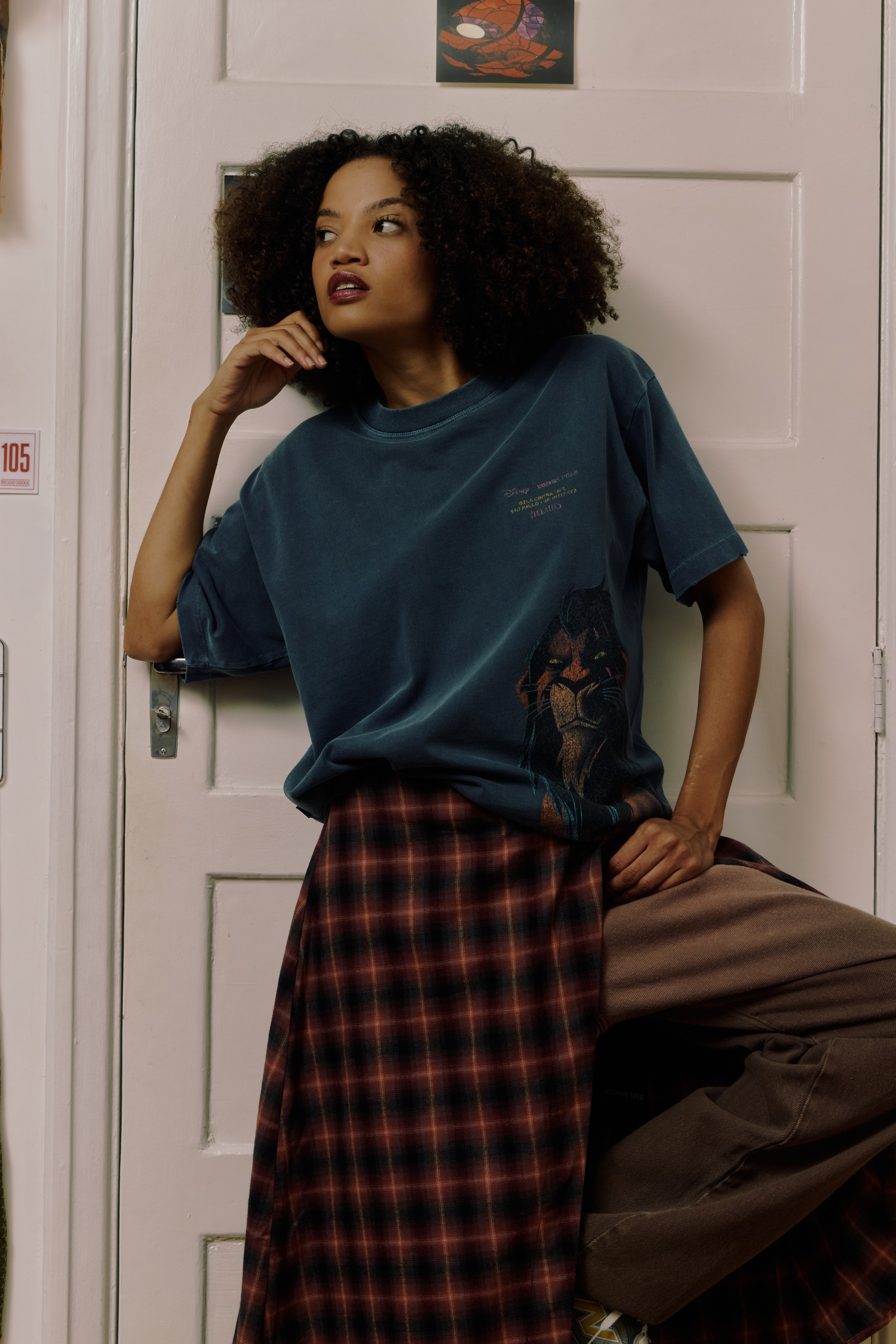 Mulher jovem de pele clara e cabelo cacheado escuro, vestindo camiseta azul-petróleo com estampa do Scar e saia xadrez vermelha e preta sobre calça marrom, posa encostada em uma porta branca com o número 105