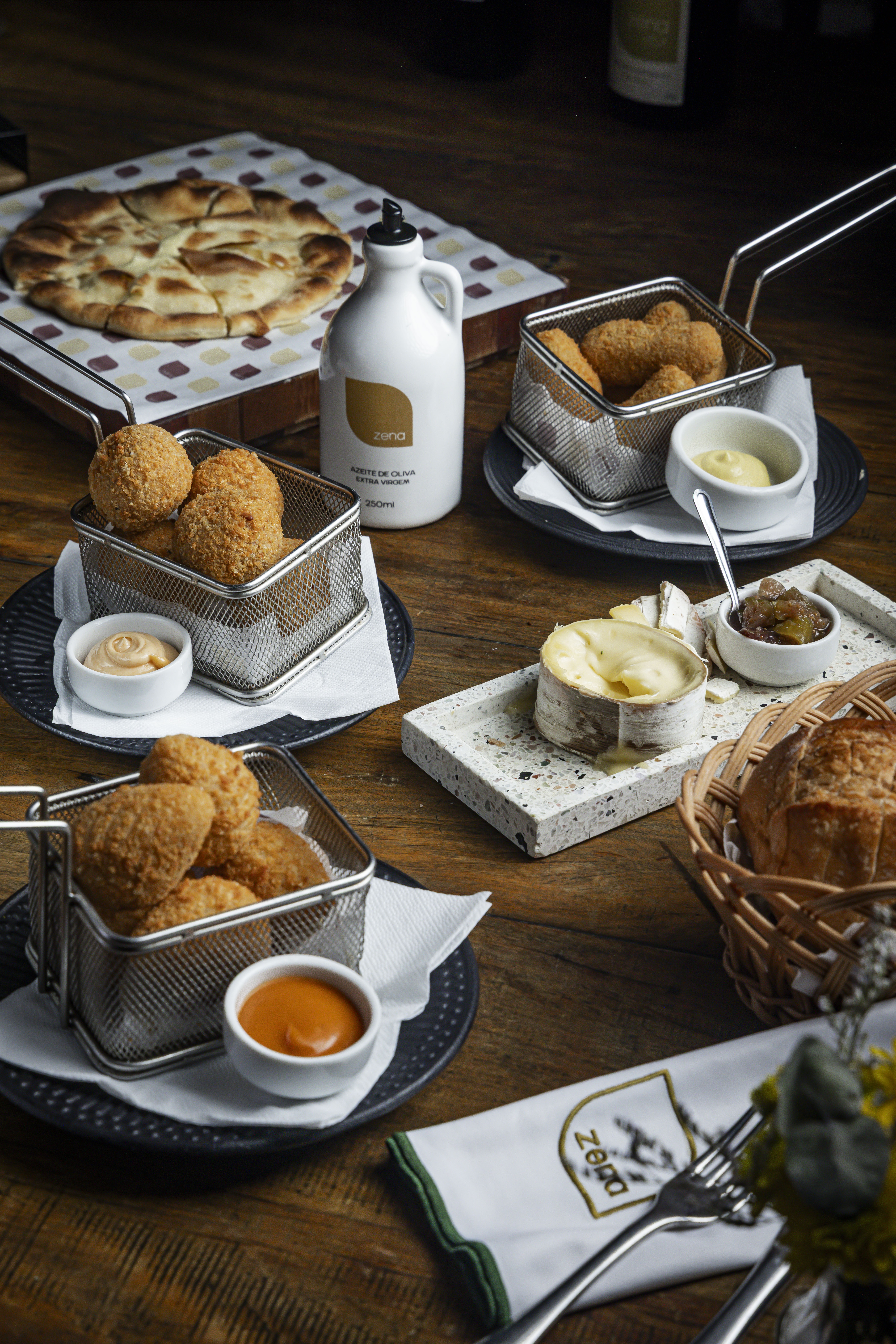 Mesa de madeira com diversos pratos: pão sírio, porções de bolinhos fritos em cestas de metal com molhos, queijo brie com geleia e pães em cesta de vime. Uma garrafa branca de azeite está no centro