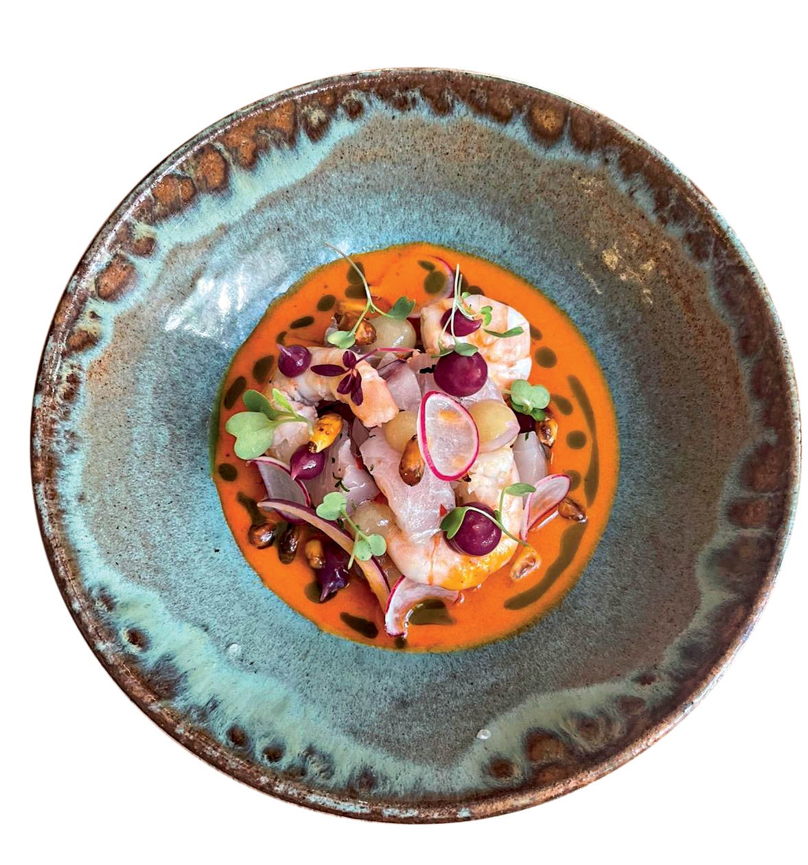 Ceviche colorido em um prato rústico azul e marrom, com camarões, peixe branco, rabanetes fatiados, uvas roxas e folhas verdes, sobre um molho laranja vibrante