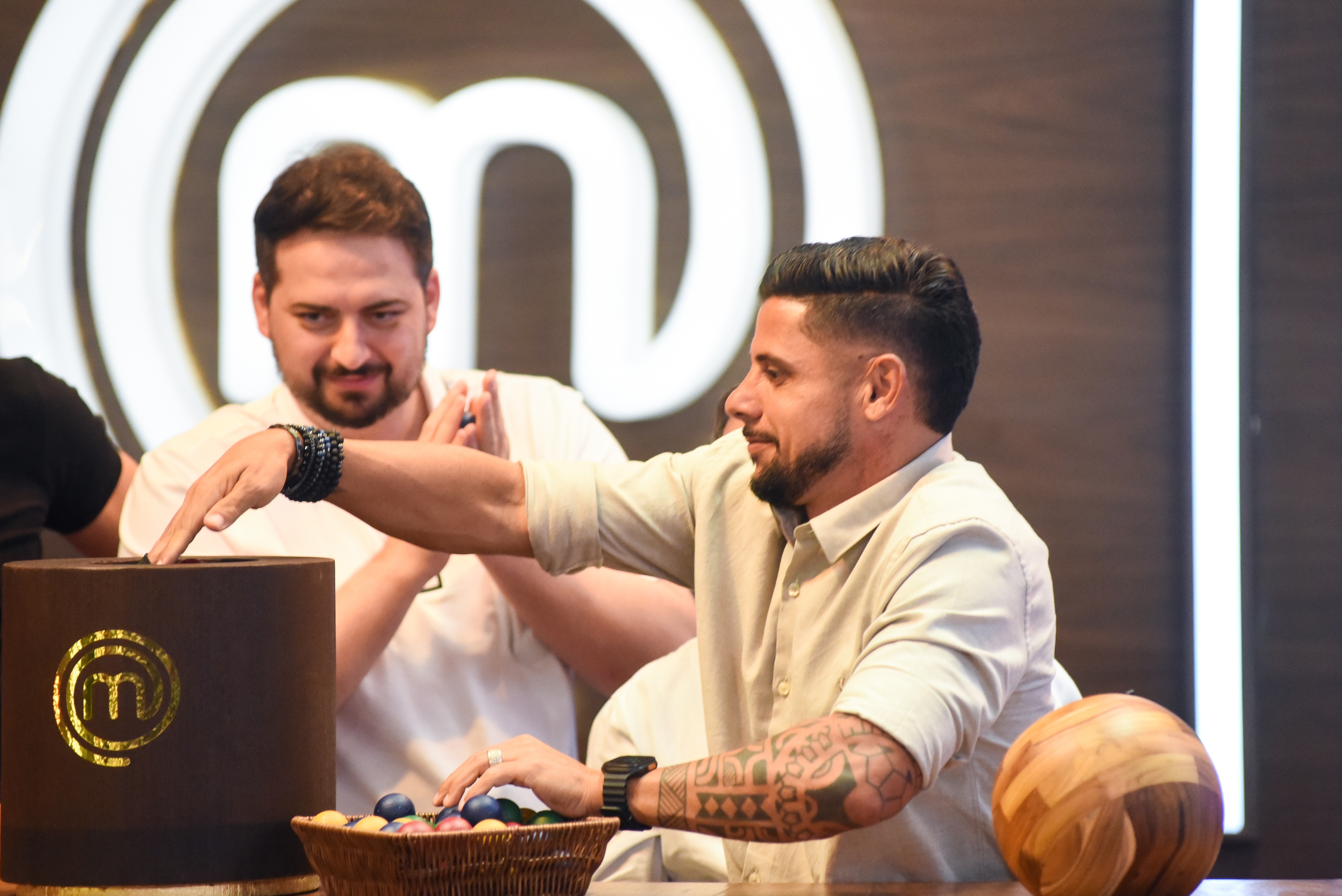 Dois homens em um evento. Um homem barbudo de camisa bege e braços tatuados pega algo de uma cesta com ovos coloridos. Outro homem barbudo de camisa branca sorri ao fundo, com a mão sobre uma caixa marrom com o logo 