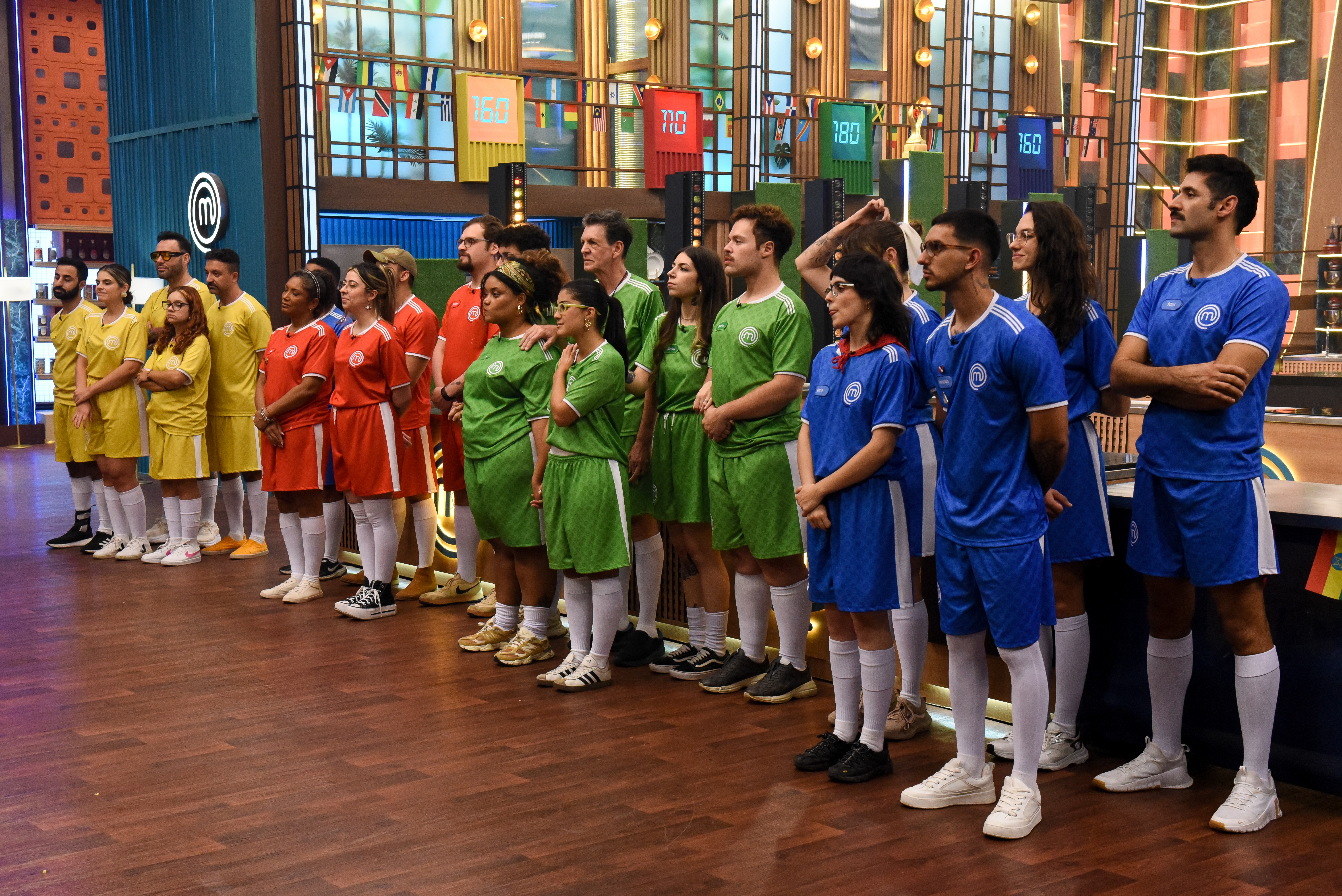 Grupo de 20 pessoas, homens e mulheres, em pé, alinhadas em um estúdio de TV. Usam uniformes esportivos de futebol nas cores azul, verde, vermelho e amarelo, com meias brancas e tênis. Ao fundo, um cenário com painéis coloridos e luzes.
