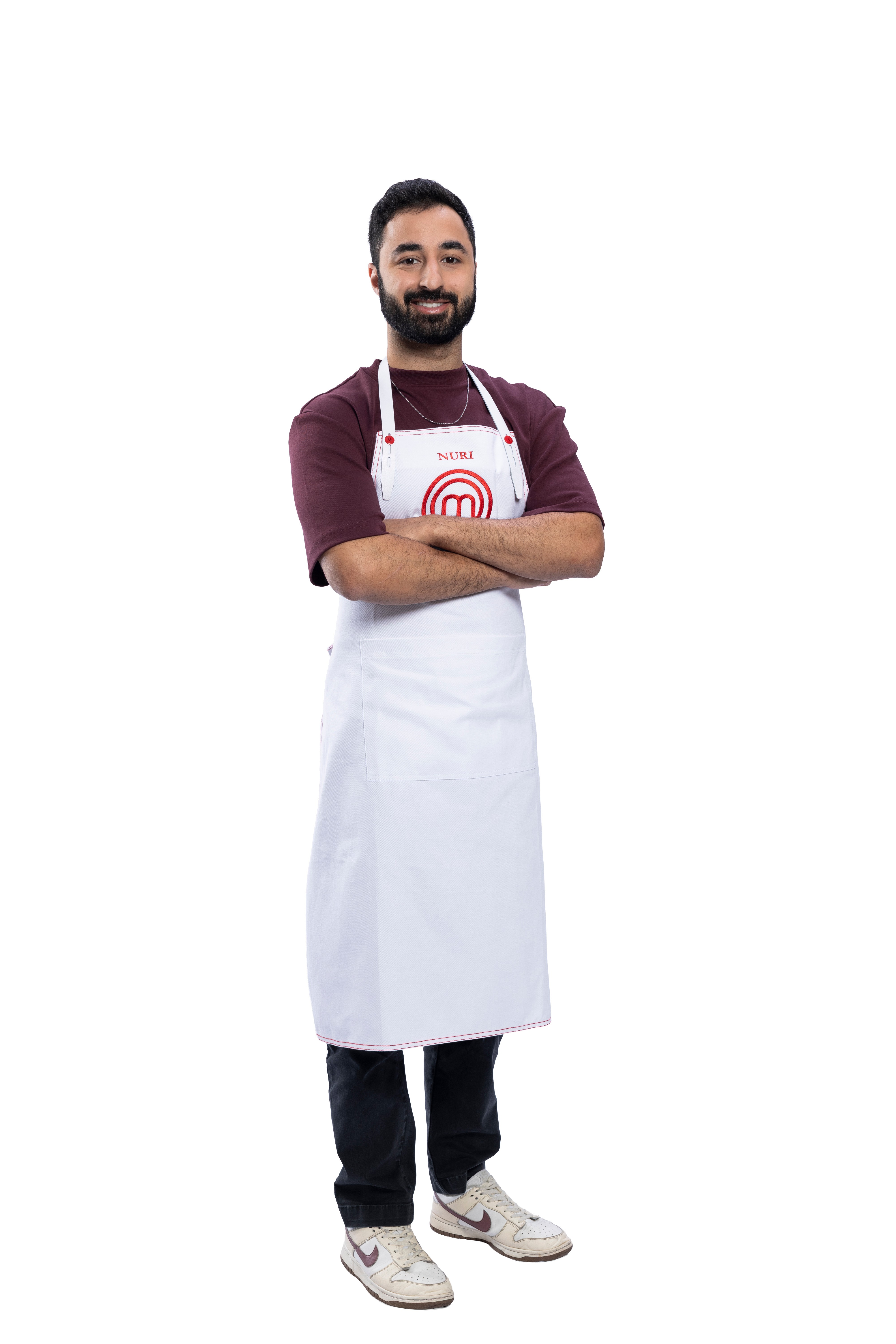 Homem barbudo, de pele morena clara, sorrindo, usando camiseta vinho e avental branco com o nome 