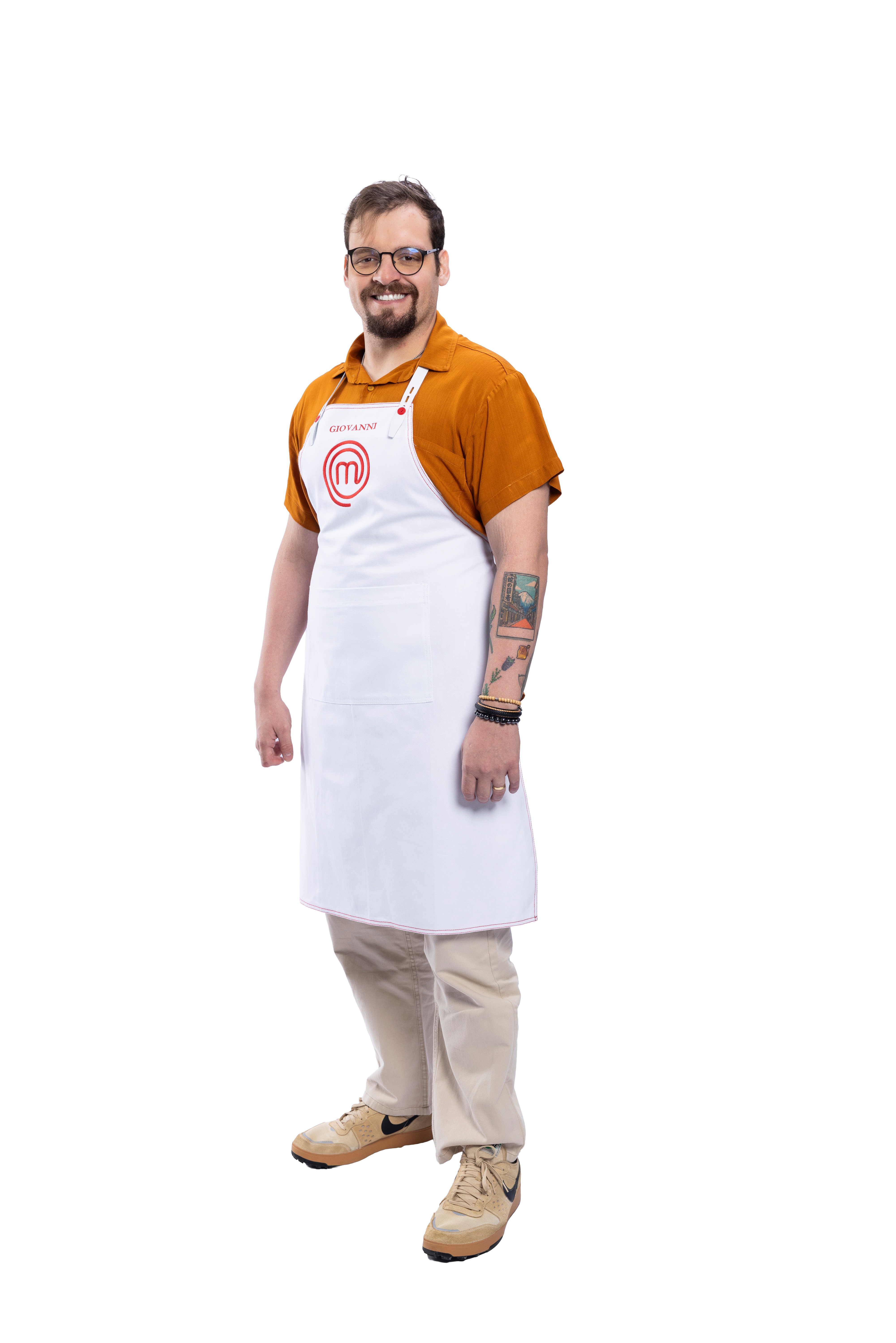 Homem barbudo, de óculos, sorrindo, vestindo camisa laranja, avental branco do MasterChef com o nome 