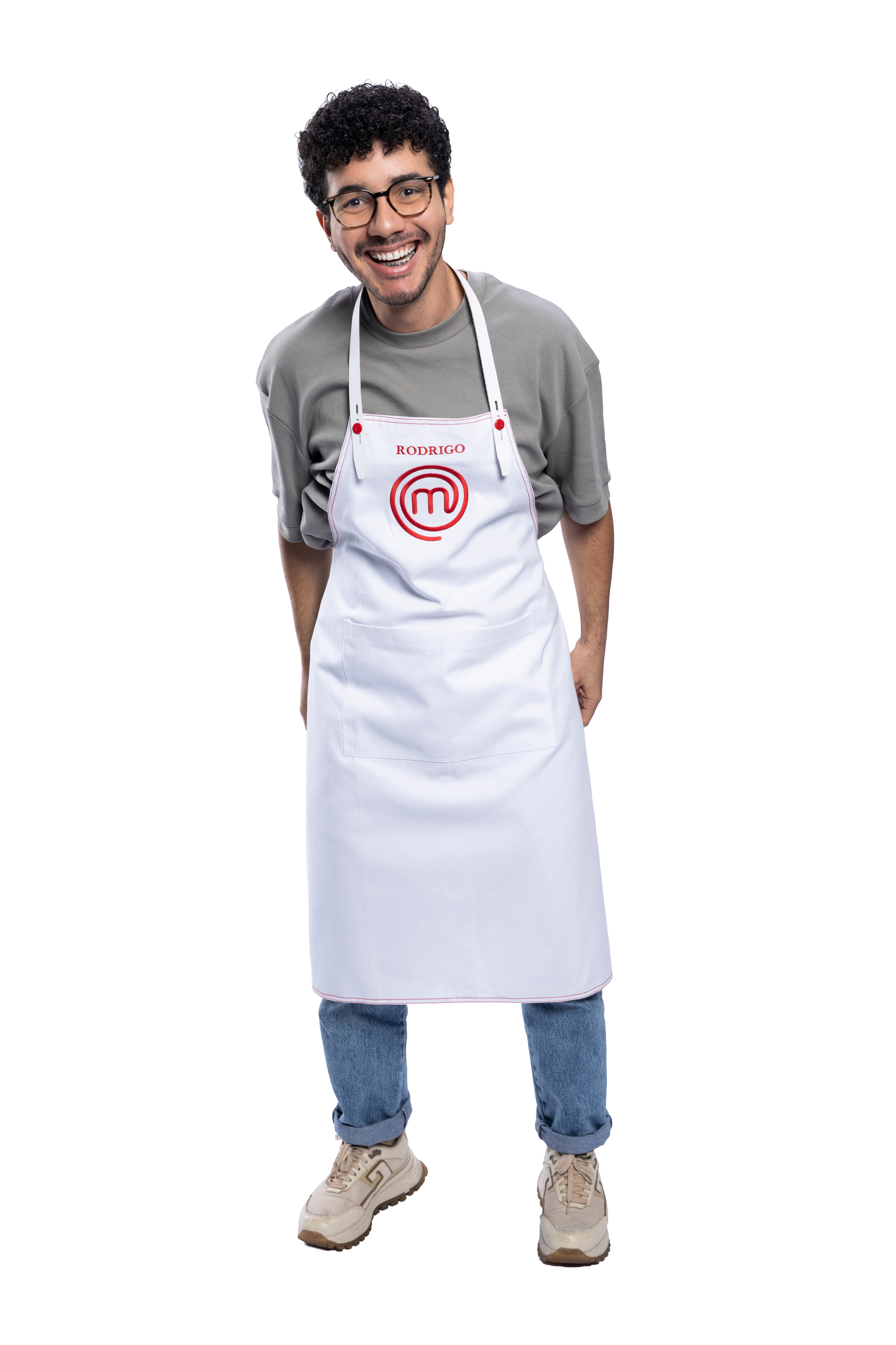 Homem jovem de pele clara, cabelo crespo escuro e óculos, sorrindo, vestindo camiseta cinza, jeans e tênis bege, com um avental branco do MasterChef escrito 