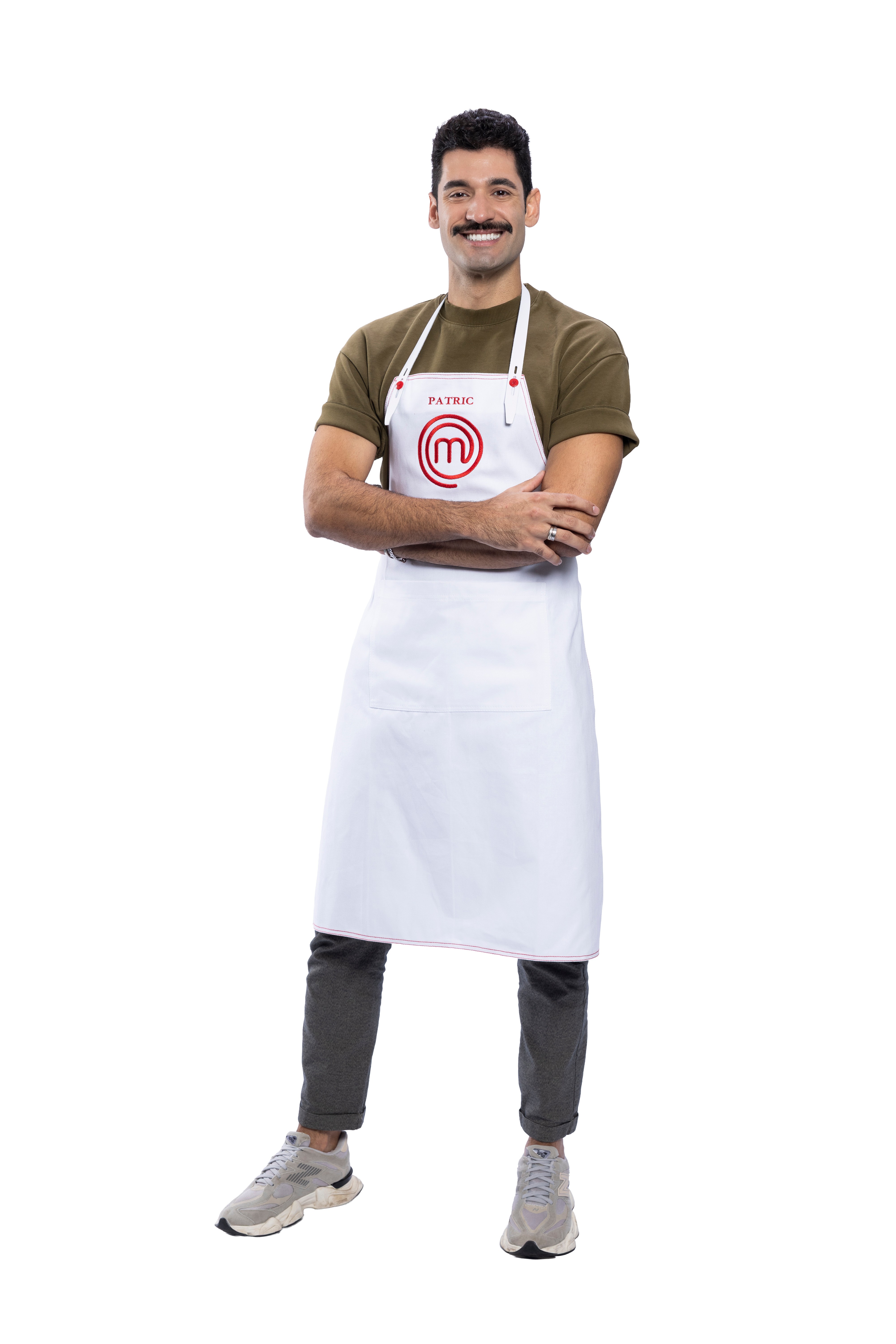 Homem sorridente com bigode, cabelo escuro e barba rala, vestindo camiseta verde-oliva, calça cinza e tênis claros. Ele usa um avental branco com o nome 