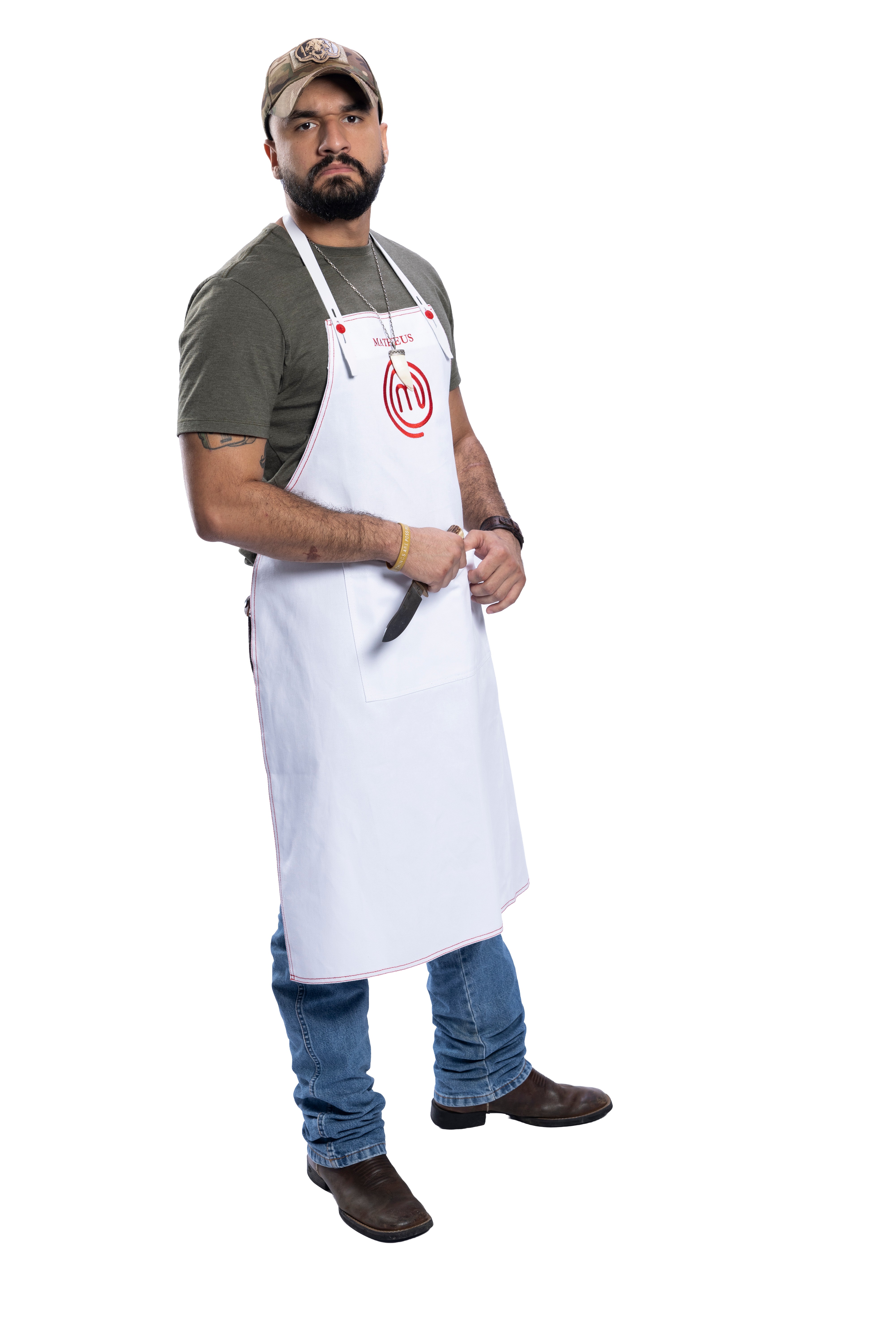 Homem barbudo, de boné camuflado, camiseta verde-oliva e avental branco com logo MasterChef, segurando uma faca, jeans e botas marrons, em fundo branco