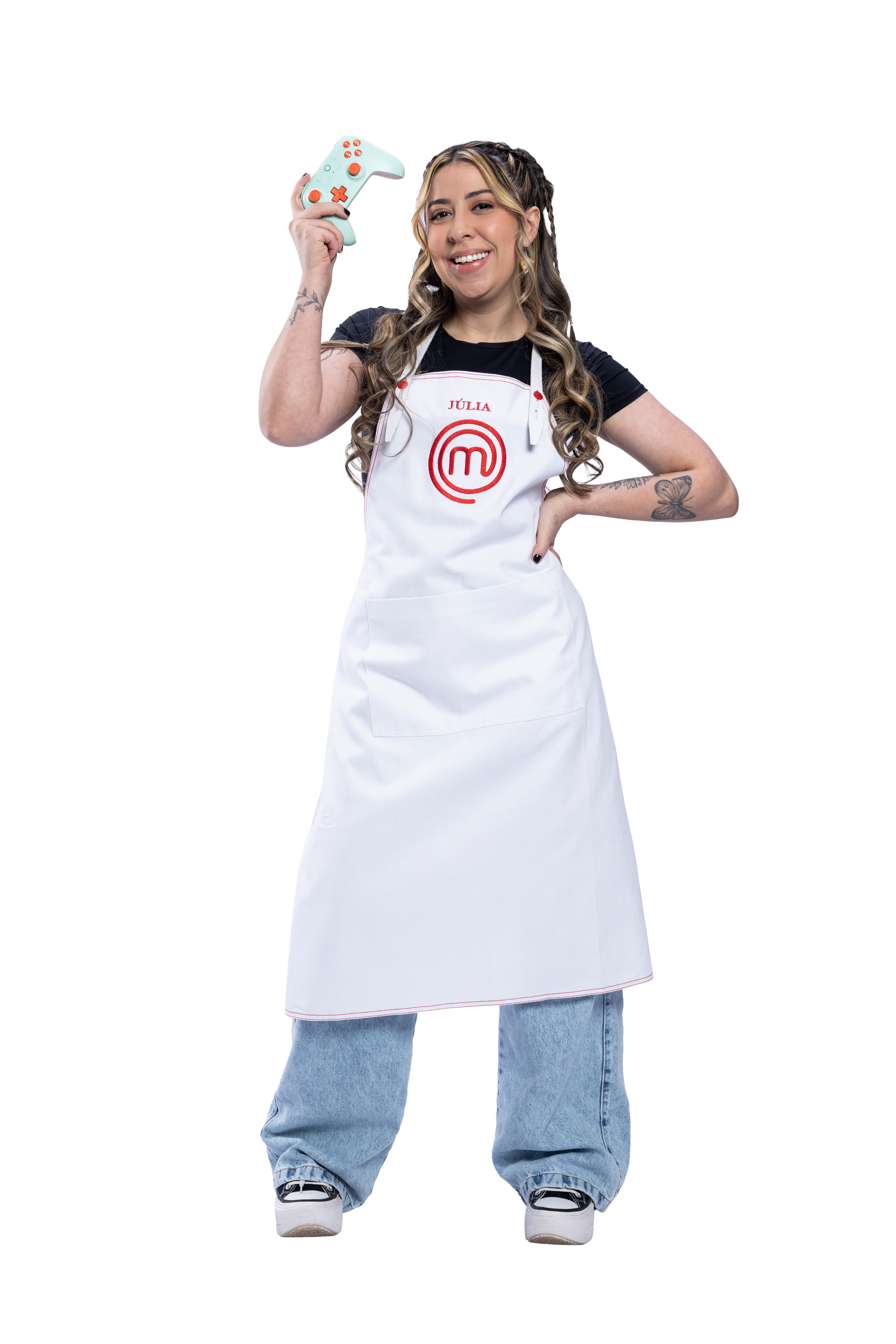 Júlia, participante do MasterChef, sorri e segura um controle de videogame verde-água com detalhes laranja, vestindo avental branco com seu nome e o logo do programa, camiseta preta e calça jeans larga